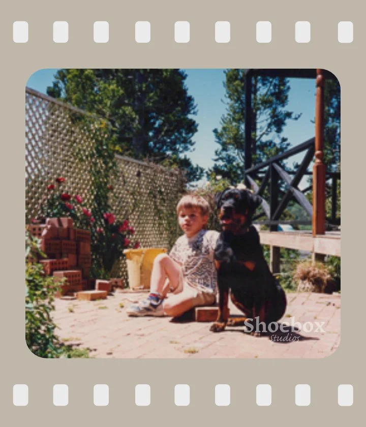 A young girl sitting on a brick patio with a big black and tan dog beside her in a backyard, with trees, a lattice fence, and a small staircase in the background.
