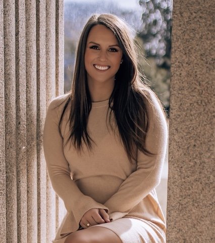 A young woman with long dark hair, smiling, wearing a beige sweater, sitting outdoors near stone columns with a blurred background of trees and sky.