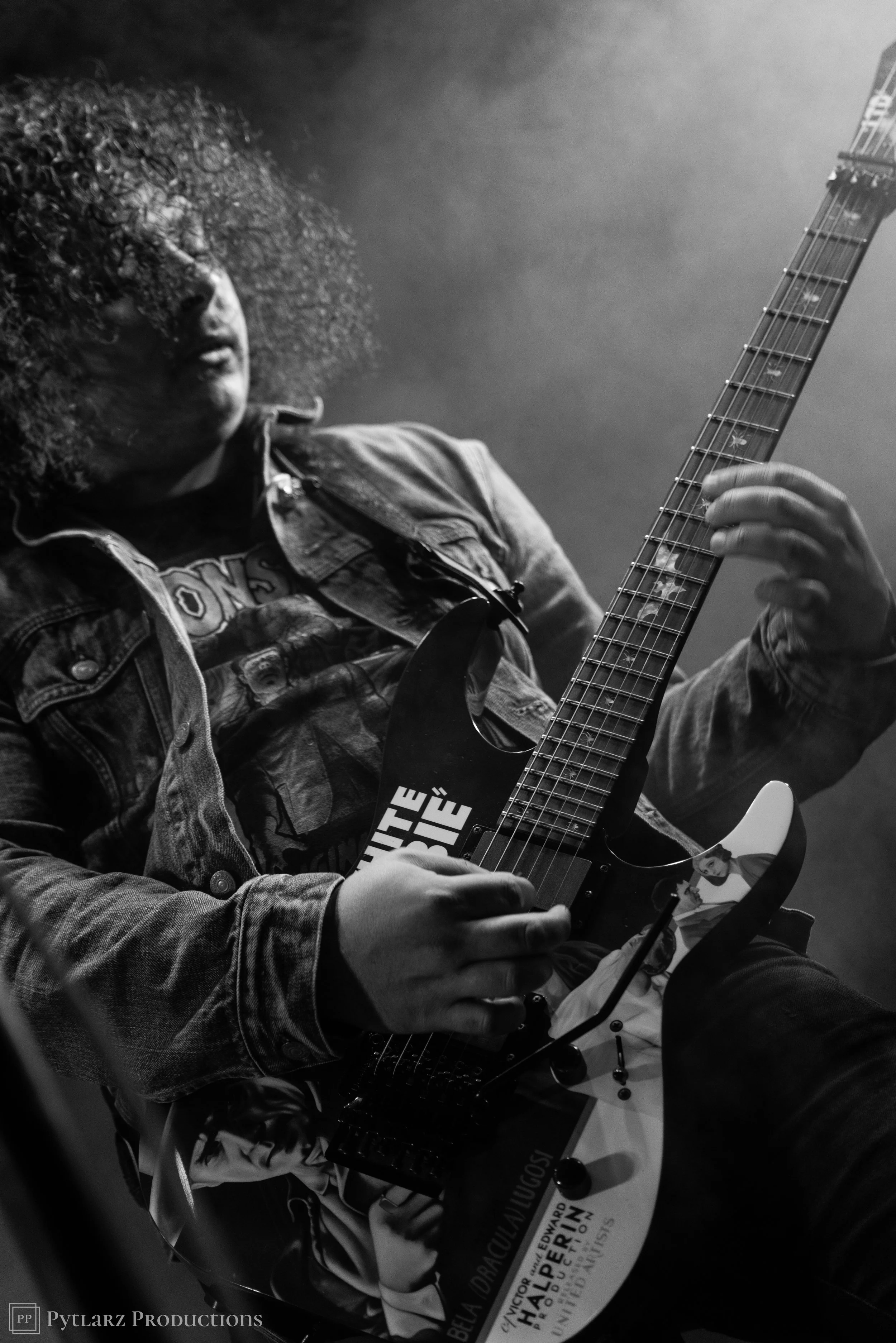 A black and white photo of a person with curly hair playing an electric guitar, wearing a denim jacket and a t-shirt with text and images, with a pin-up girl illustration on the guitar. The person's face is partially obscured by hair and the backgrou