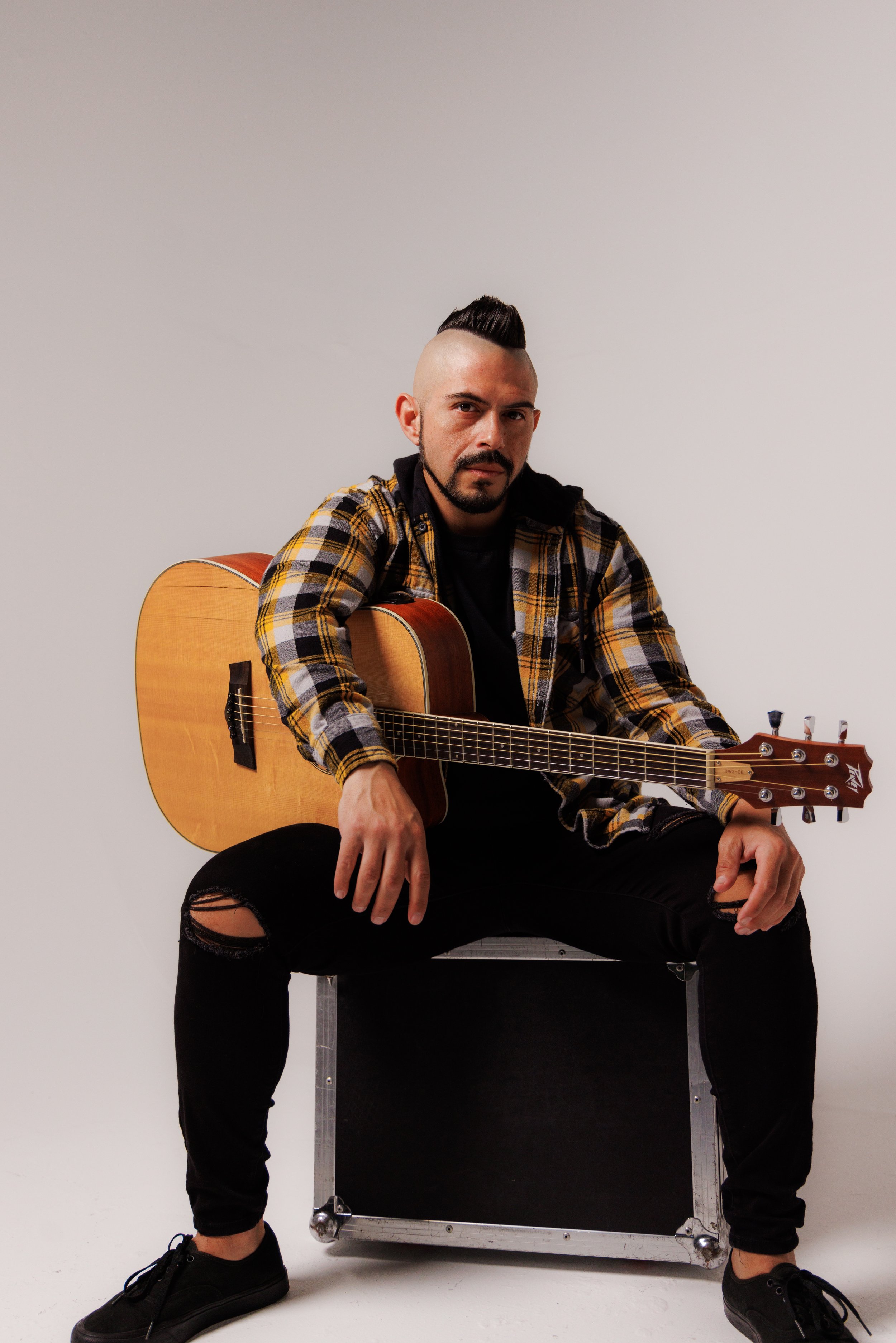 A man with a mohawk hairstyle, beard, and tattoos is sitting on a black box, holding an acoustic guitar, wearing a yellow and black plaid shirt, ripped black jeans, and black sneakers, against a plain white background.