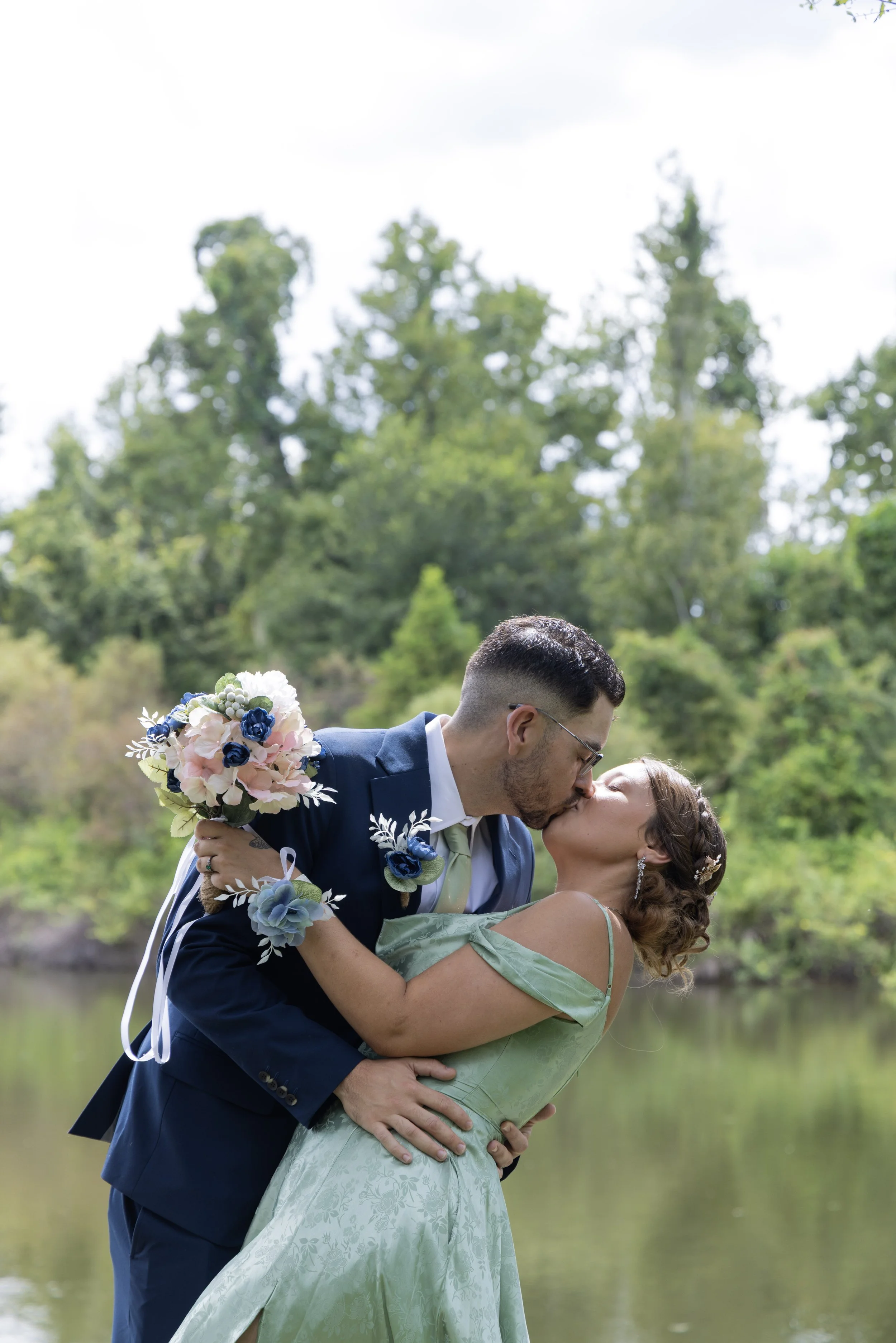 A couple dressed in wedding attire sharing a kiss outdoors near a river with green trees in the background, the groom holding a bouquet of pink, white, and blue flowers.
