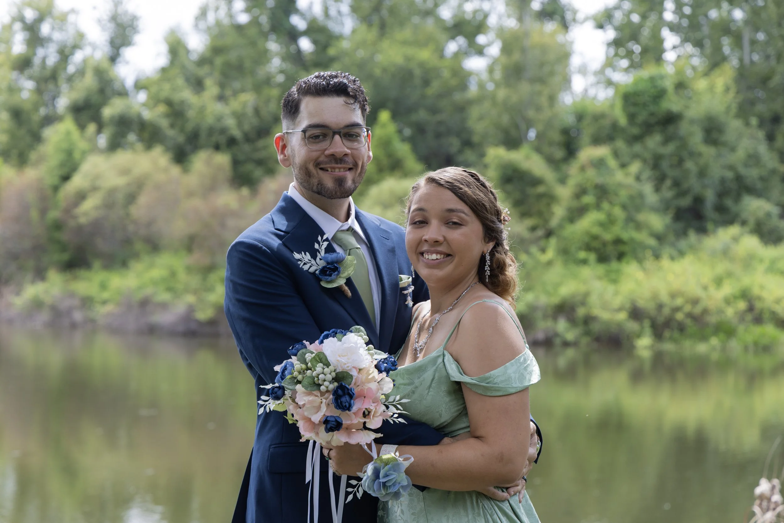 A smiling couple in wedding attire standing outdoors by a body of water with lush green trees in the background, holding a bouquet of pink, white, and blue flowers.