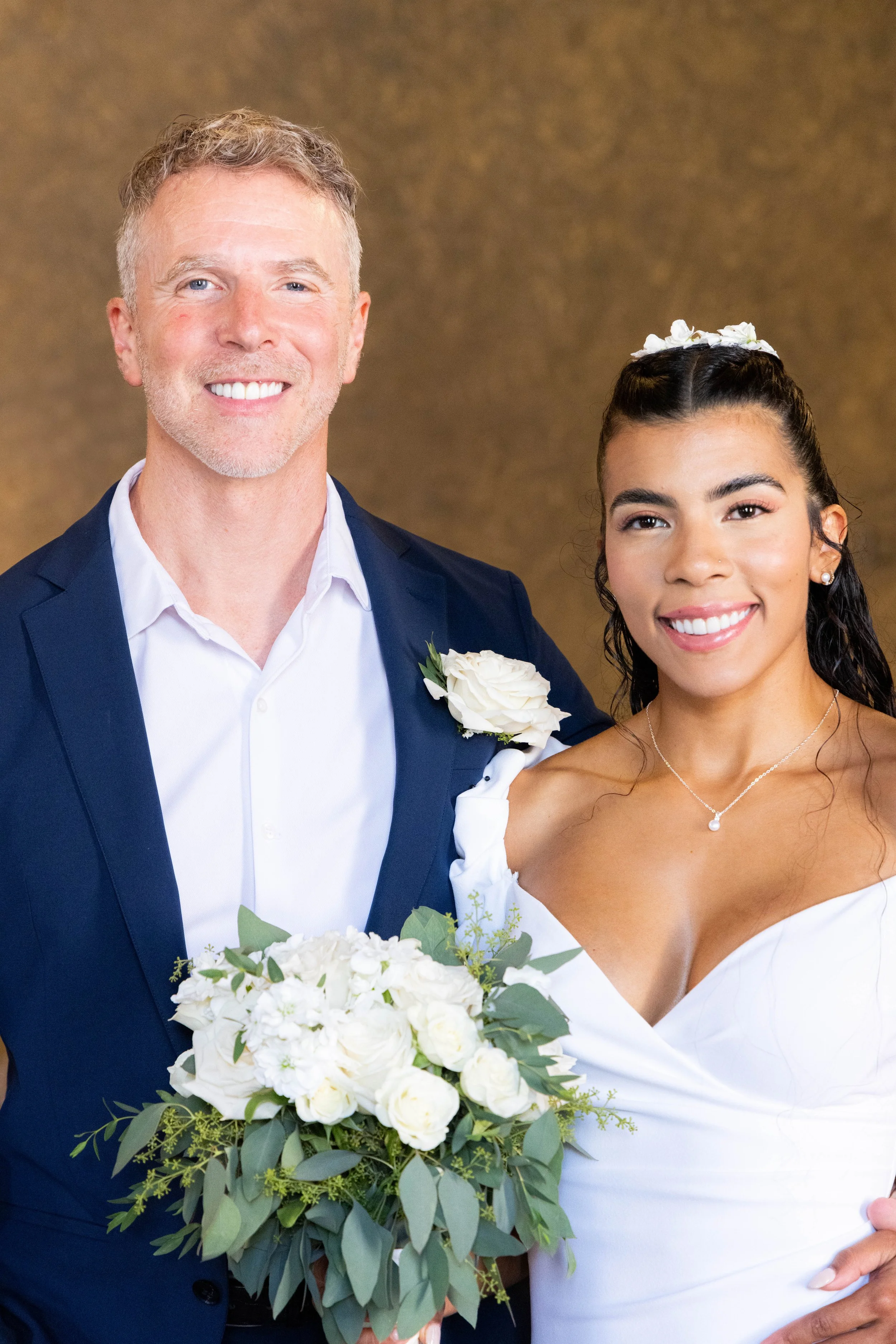 A smiling man in a navy suit and white shirt standing next to a smiling woman in a white dress. The man is holding a bouquet of white flowers and greenery. The woman has dark hair styled in waves, with white flowers in her hair and a pearl necklace. 