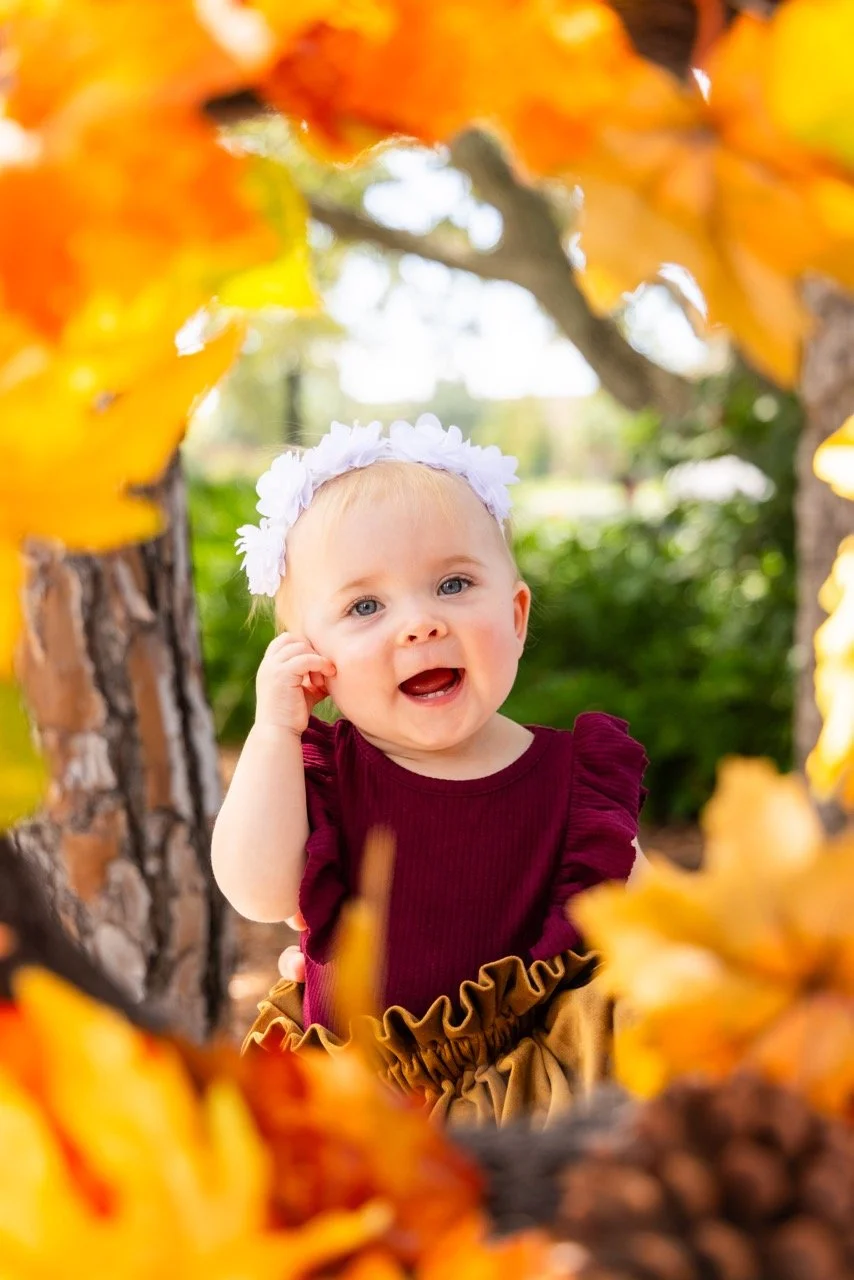 A smiling toddler girl with a white flower headband, wearing a maroon top and ruffled tan skirt, looking through a frame of yellow and orange autumn leaves outdoors.