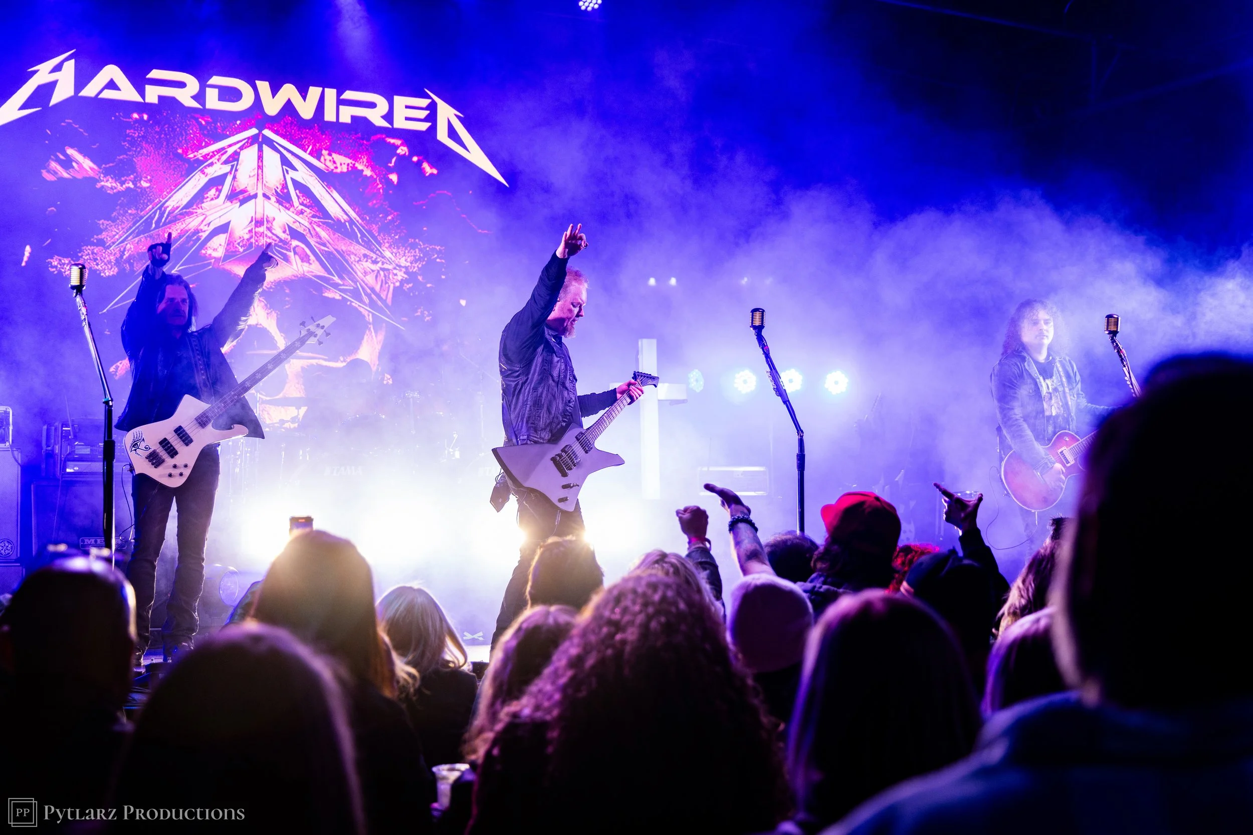 A live concert scene with band members on stage, playing guitars and singing, in front of a large screen displaying "HARDWIRE". The audience is visible at the bottom, with raised hands and heads, under colorful stage lighting and fog.