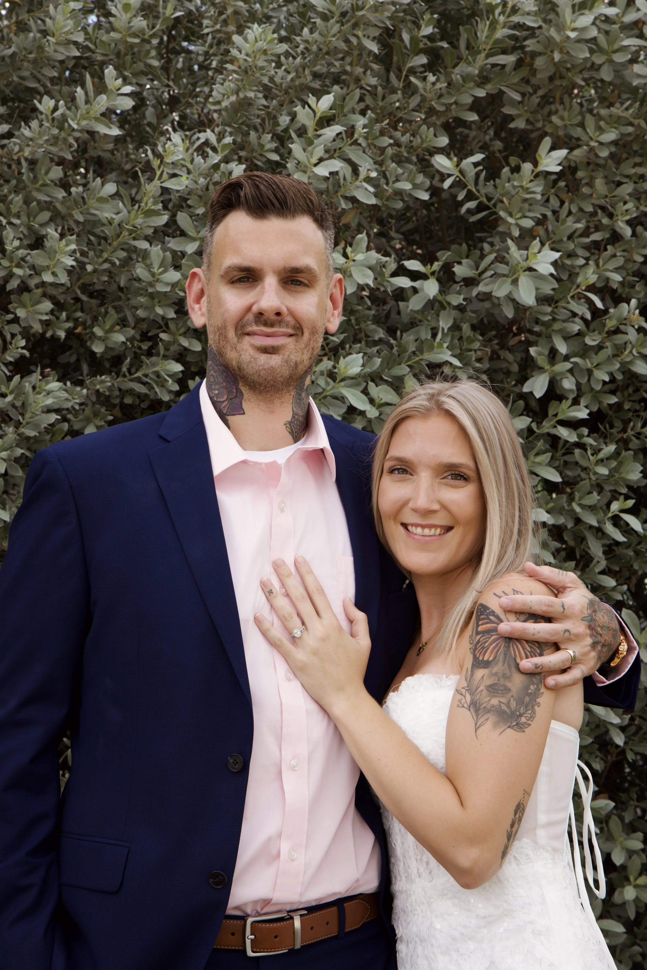 A couple dressed in wedding attire celebrating outdoors, with greenery in the background. The man is wearing a navy blue suit with a light pink shirt, and has tattoos on his neck. The woman is in a white strapless wedding dress, has tattoos on her ar