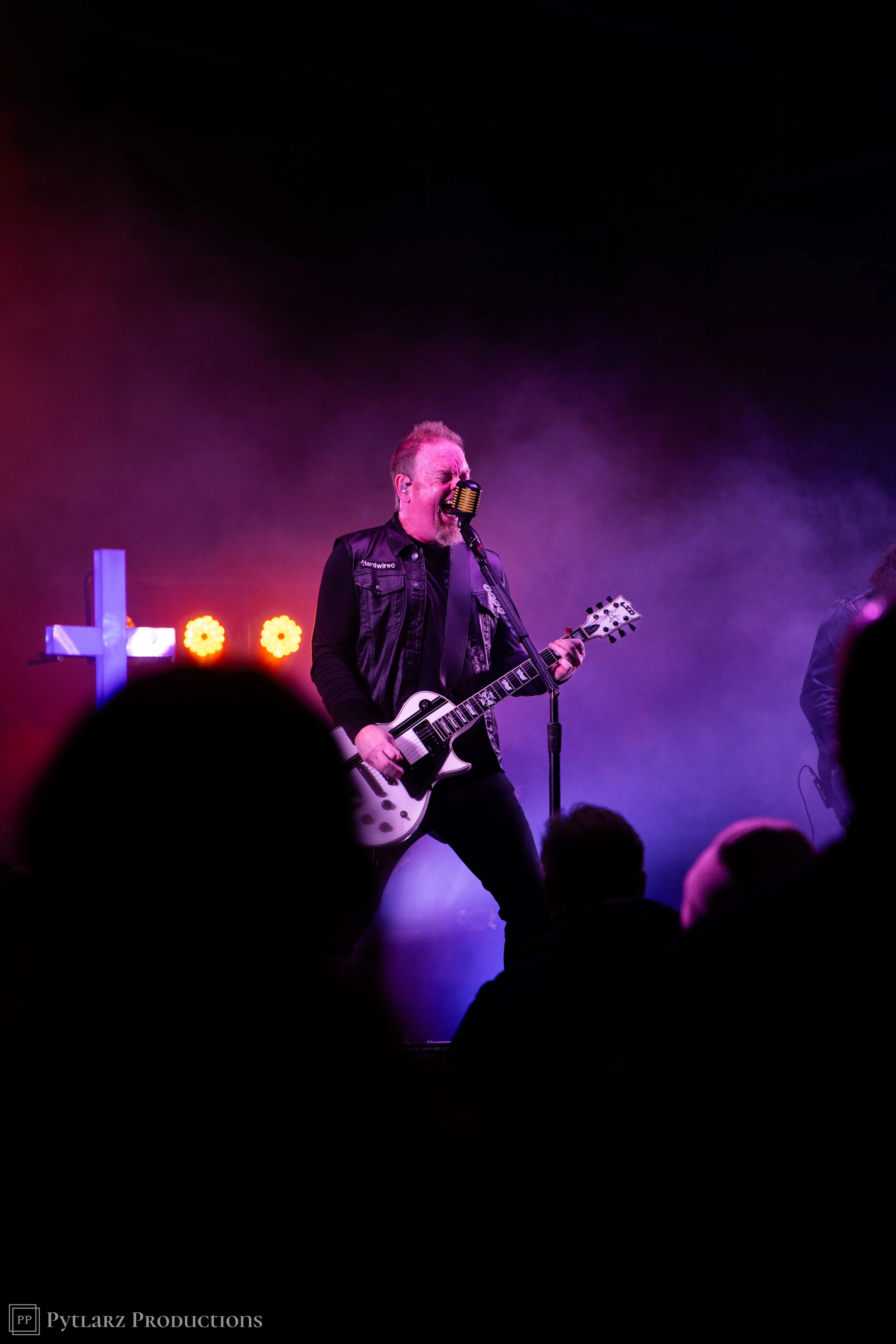 A man singing passionately into a vintage style microphone while playing an electric guitar during a live performance, illuminated by colorful stage lights.