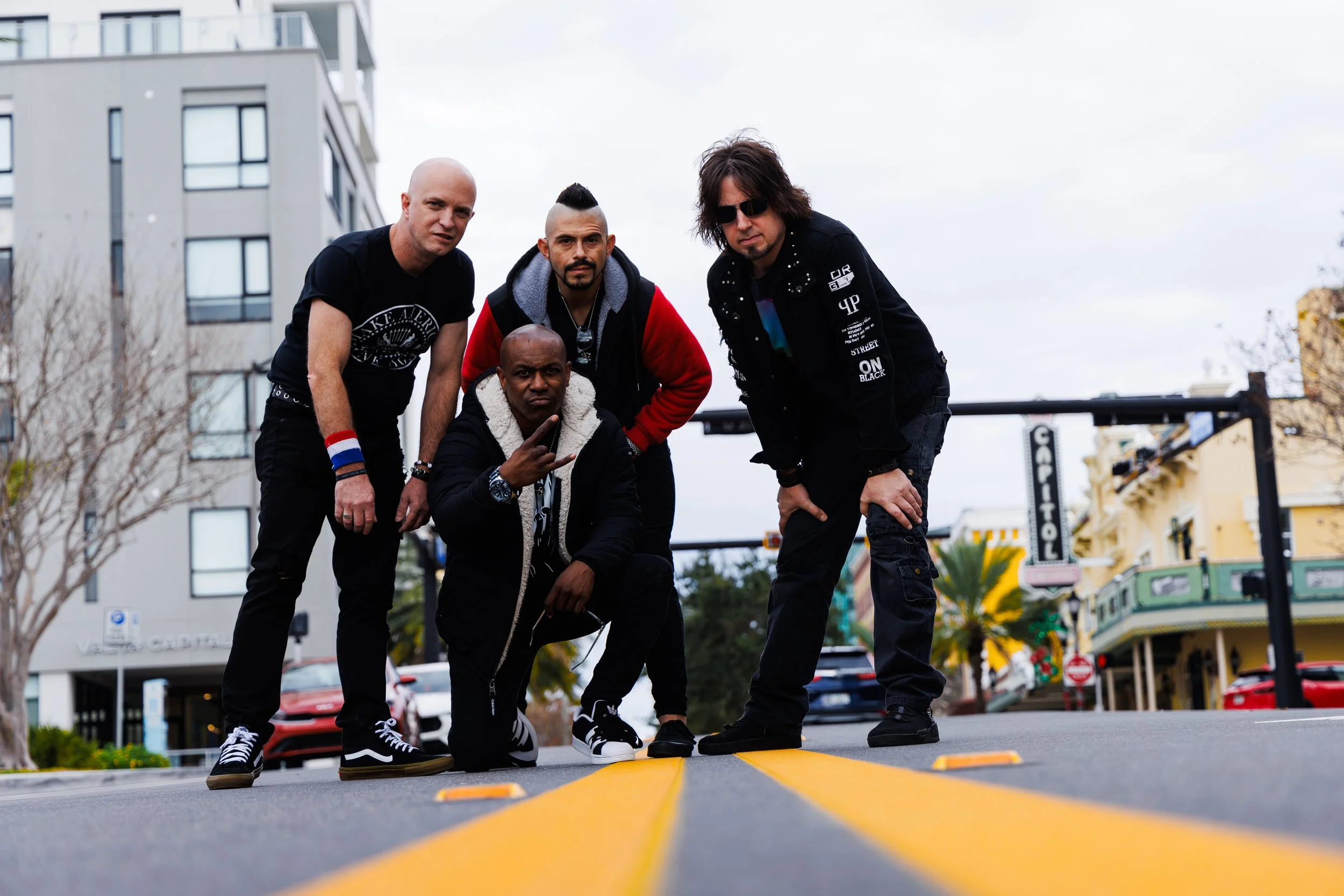 Four men posing on a city street with buildings in the background. They are squatting or kneeling on a yellow road marking.