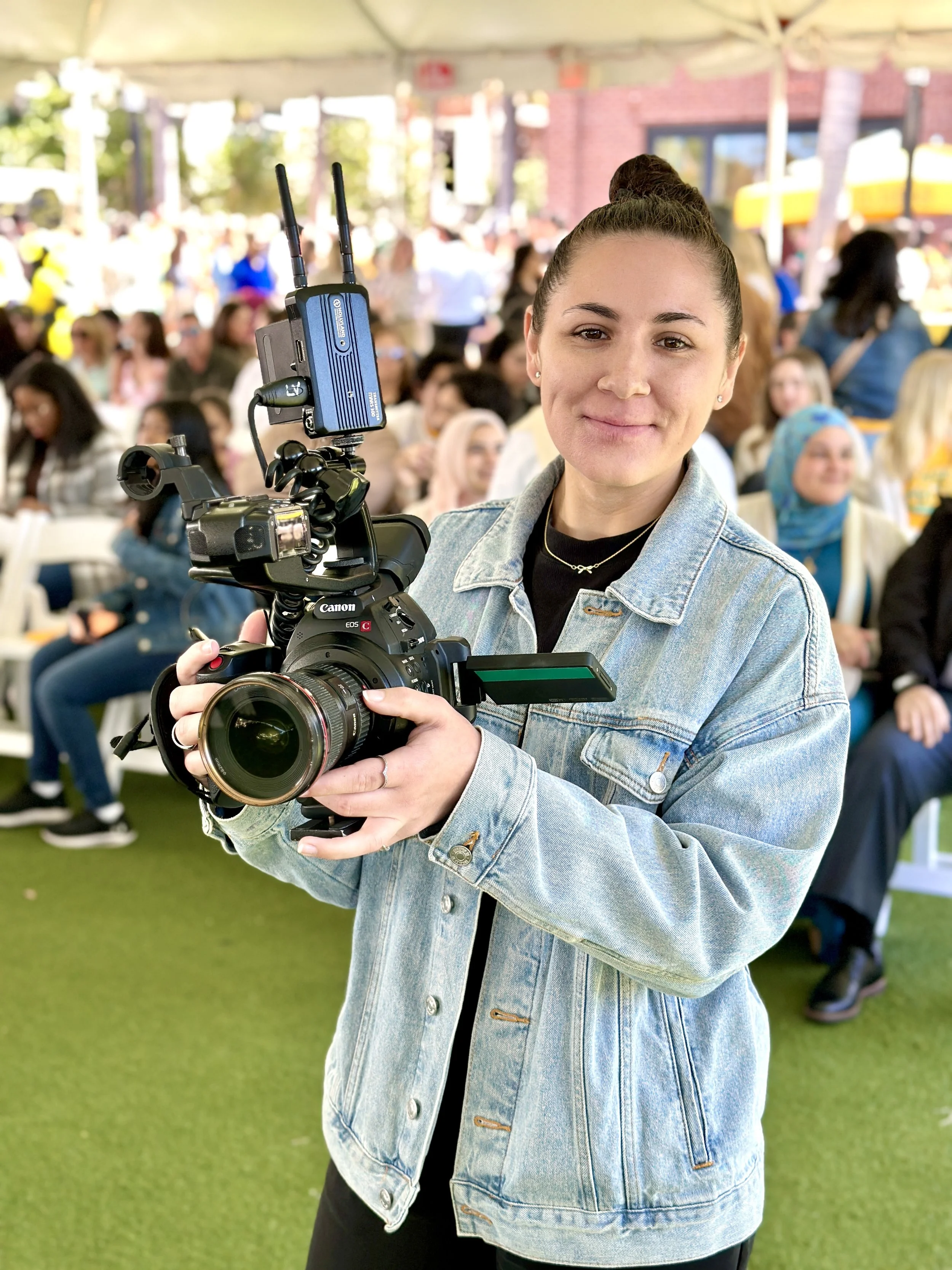 Young woman holding a professional video camera at an outdoor event under a tent with a crowd of people in the background.