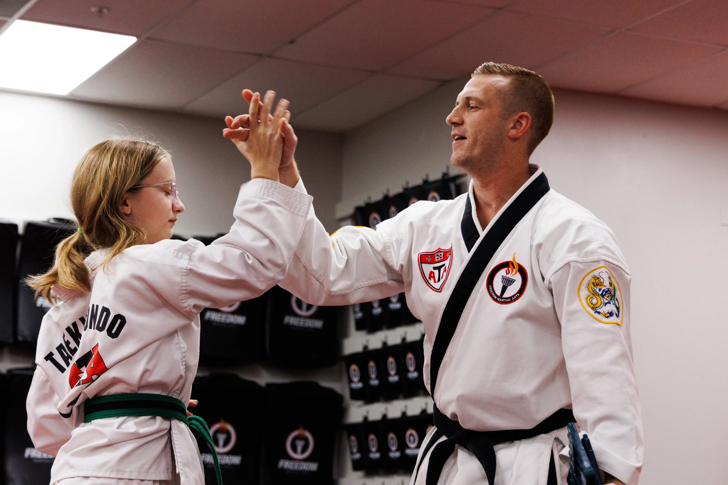 Two martial artists, one male and one female, wearing white uniforms with patches, high-five each other in a training room with black martial arts gear on a shelf in the background.