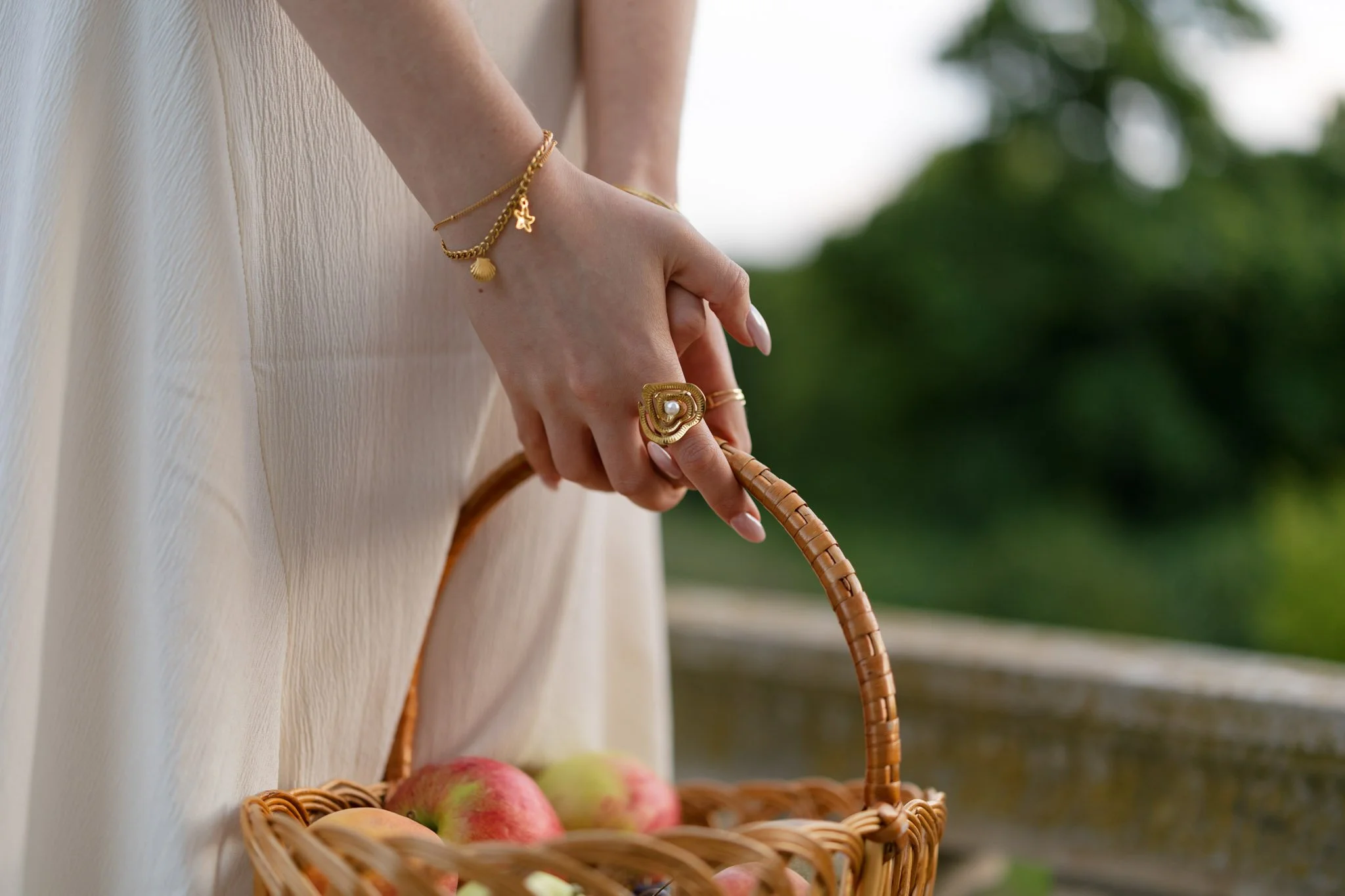 Close-up of a person's hand holding a wicker basket filled with apples. The person is wearing gold jewelry including a bracelet with shell and star charms and a large gold ring with a pearl. The background shows greenery and a blurred outdoor setting.