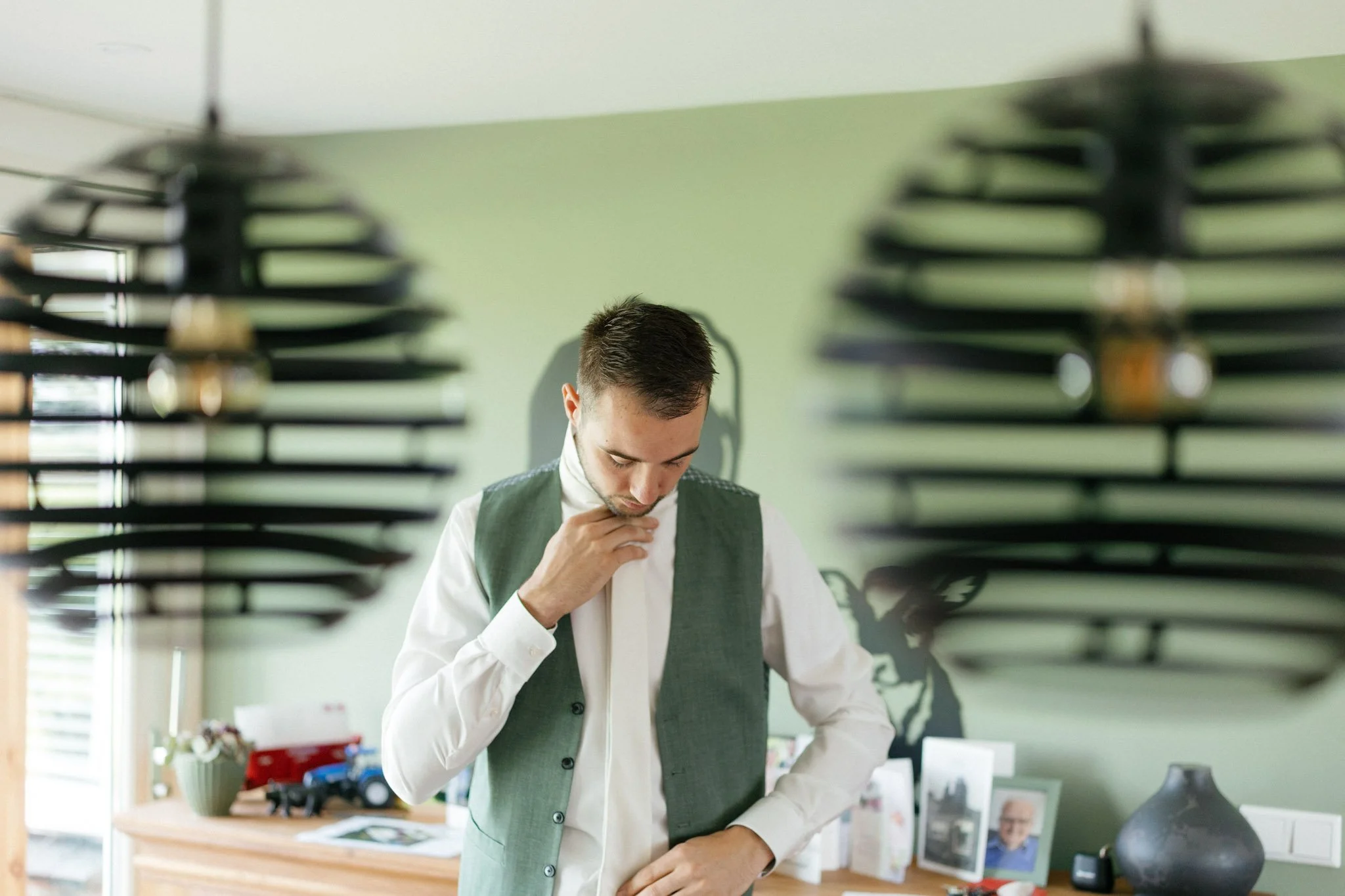 A man in a white shirt and green vest looking down thoughtfully, framed by blurry window blinds in the foreground, with a workspace and framed photograph in the background.