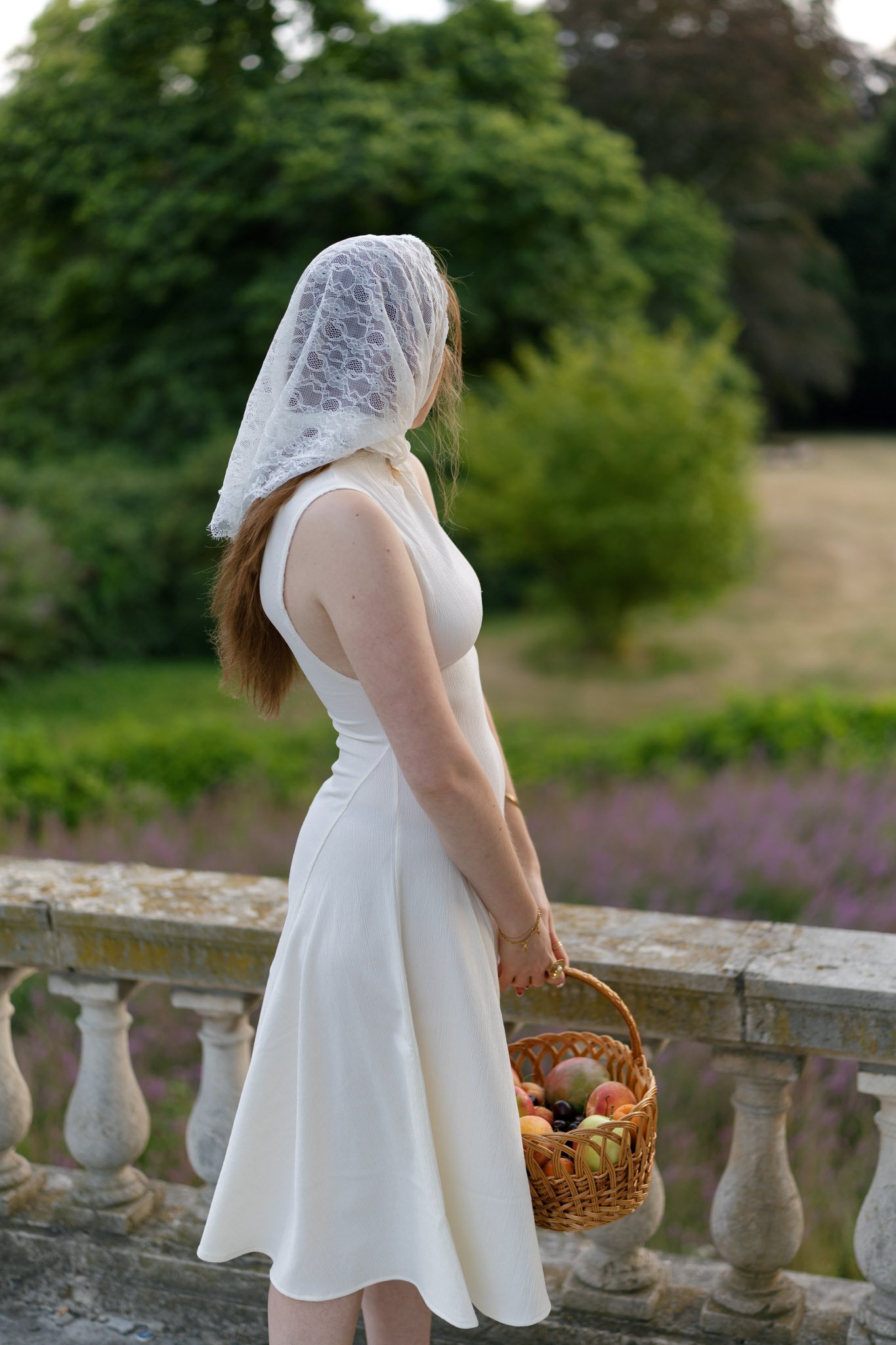 A woman wearing a white dress and lace headscarf standing by a stone balustrade outdoors, holding a wicker basket filled with apples, with green trees in the background.