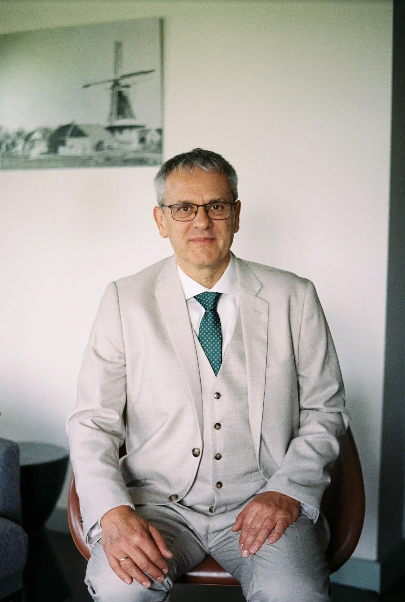 A middle-aged man wearing glasses and a light-colored suit with a green polka dot tie, sitting on a chair in a professional setting with a white wall and a black-and-white helicopter photograph in the background.
