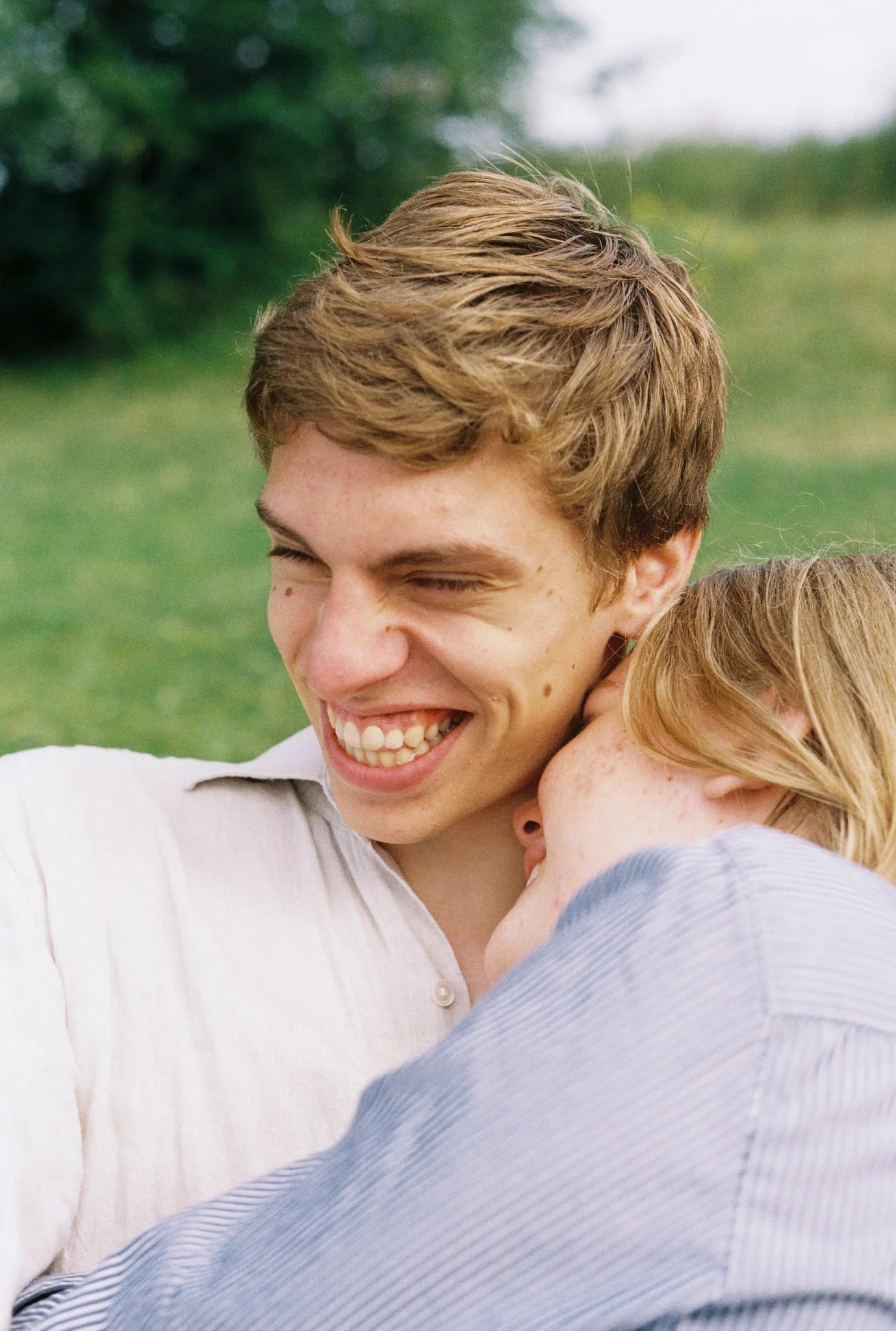 A young man with light brown hair smiling and hugging a red-haired child outdoors on a grassy field.