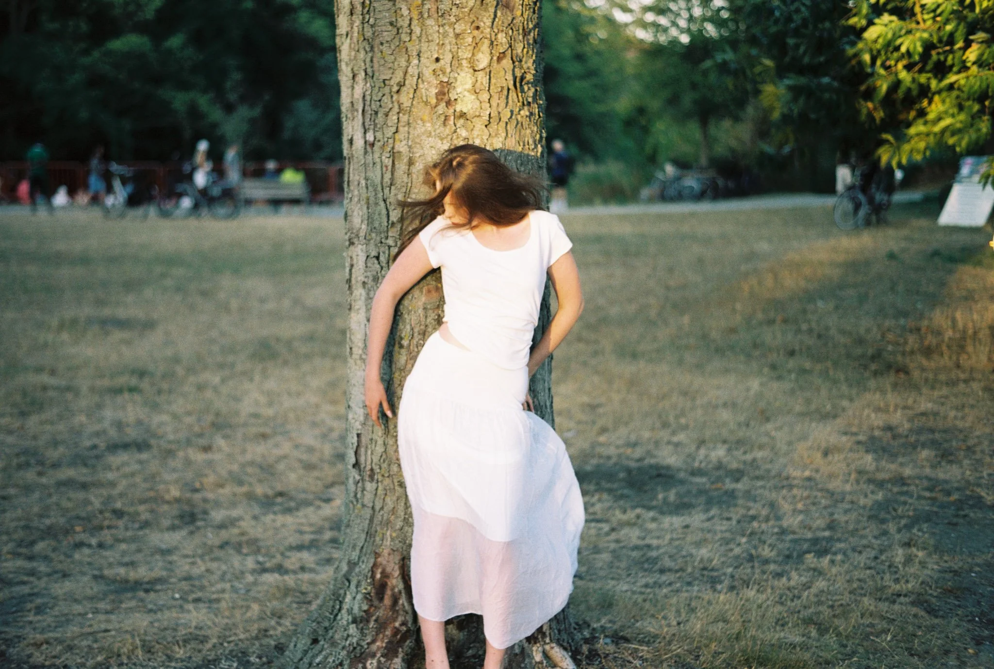 A woman in a white dress with long brown hair leaning against a tree in a park during sunset, with blurred people and greenery in the background.