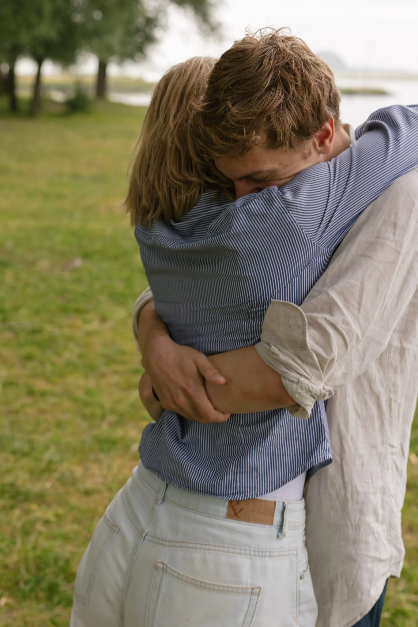 Two people hugging outdoors with trees and water in the background.