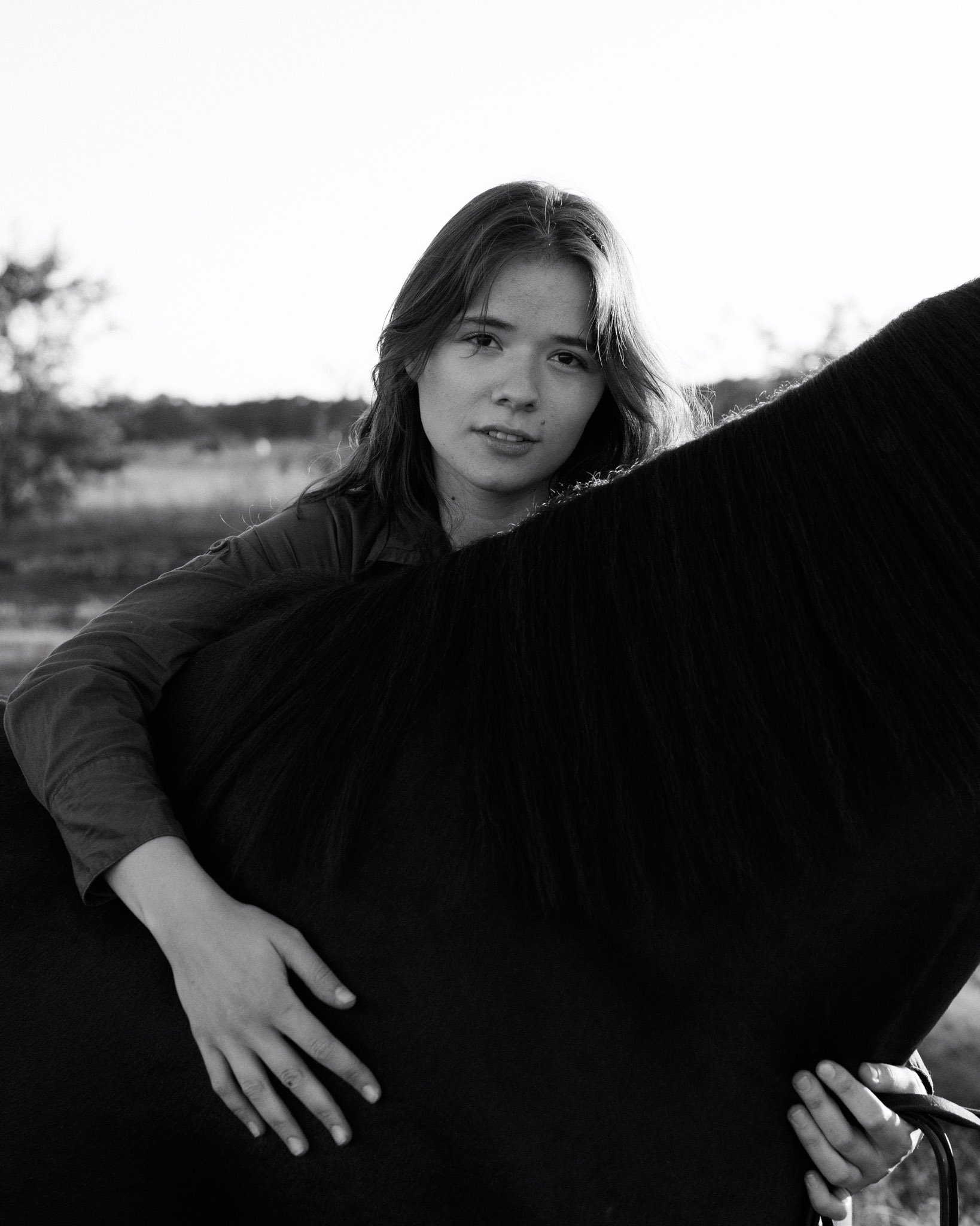 The image shows a woman with long hair holding a black horse, with the background of an outdoor field and trees, in black and white.