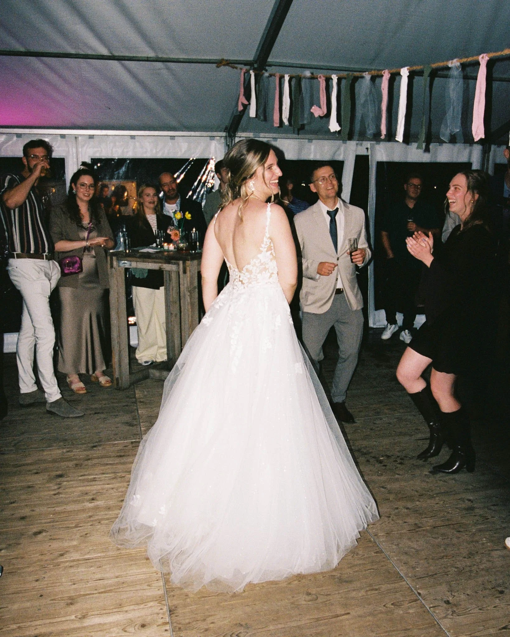 Wedding reception with woman in white dress and people around, some with drinks, inside a tent with hanging fabric decorations.