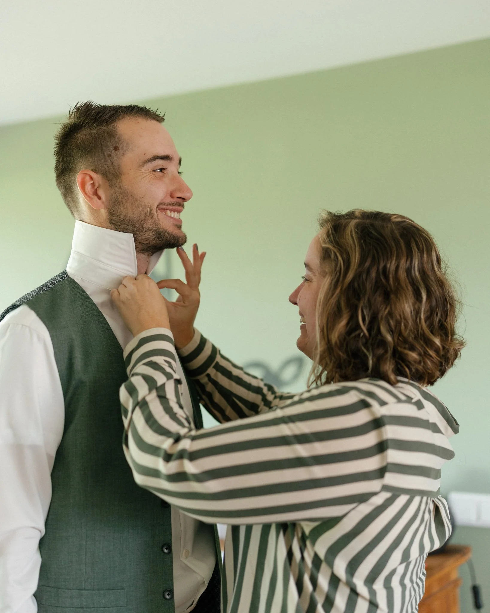 A man in a suit with a white shirt and a vest is smiling while a woman adjusts his collar. They are standing close to each other against a light green wall.