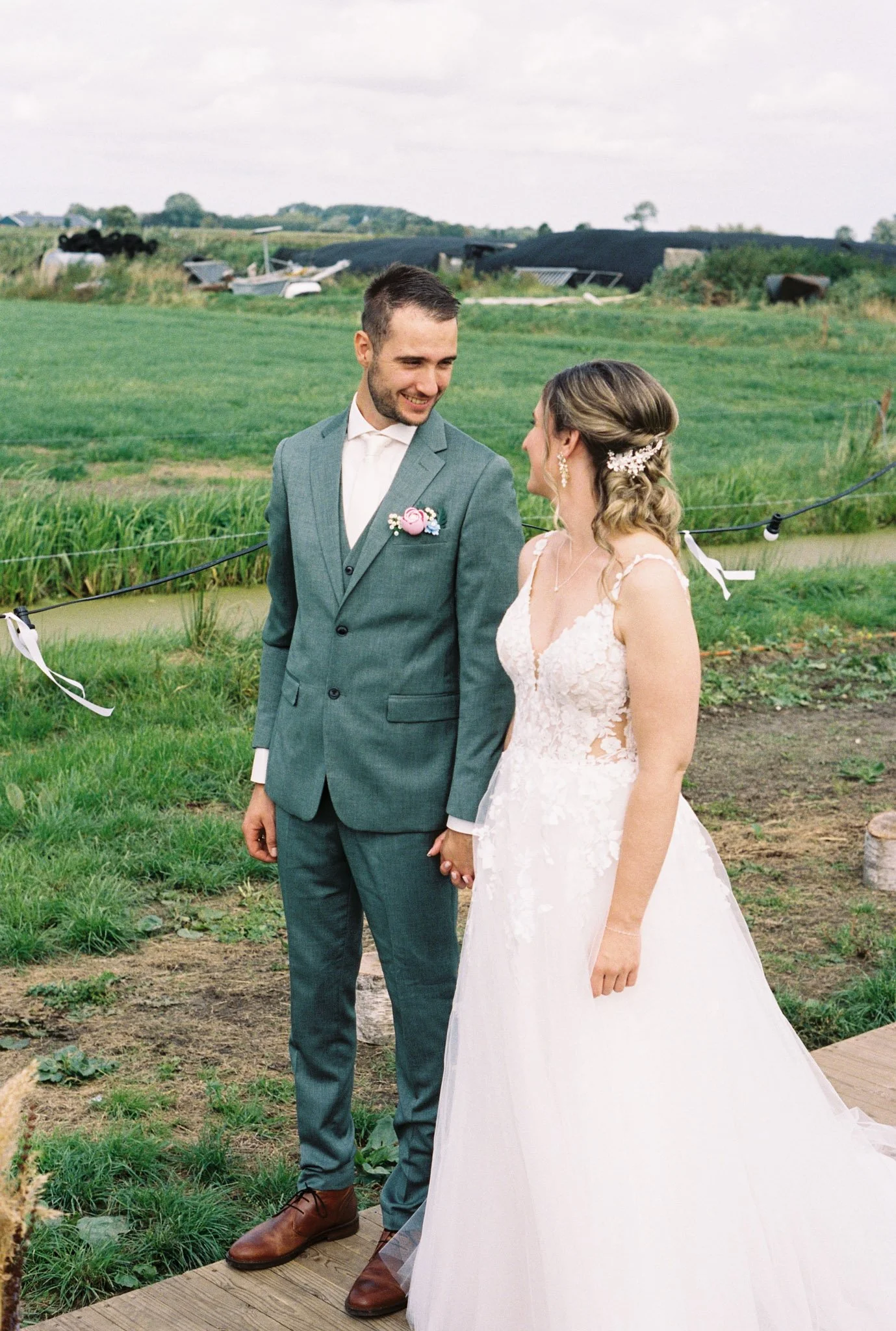 A smiling bride and groom holding hands outdoors on their wedding day, with the bride wearing a lace wedding gown and the groom in a green suit, in a rural setting with green fields and cloudy sky.