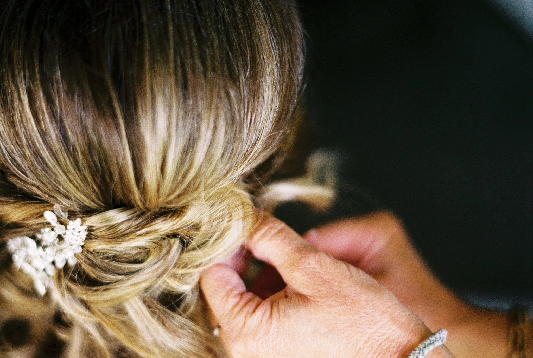 A close-up view of a bride's hair and handmade jewelry, with a person's hand adjusting her hair during a wedding ceremony.