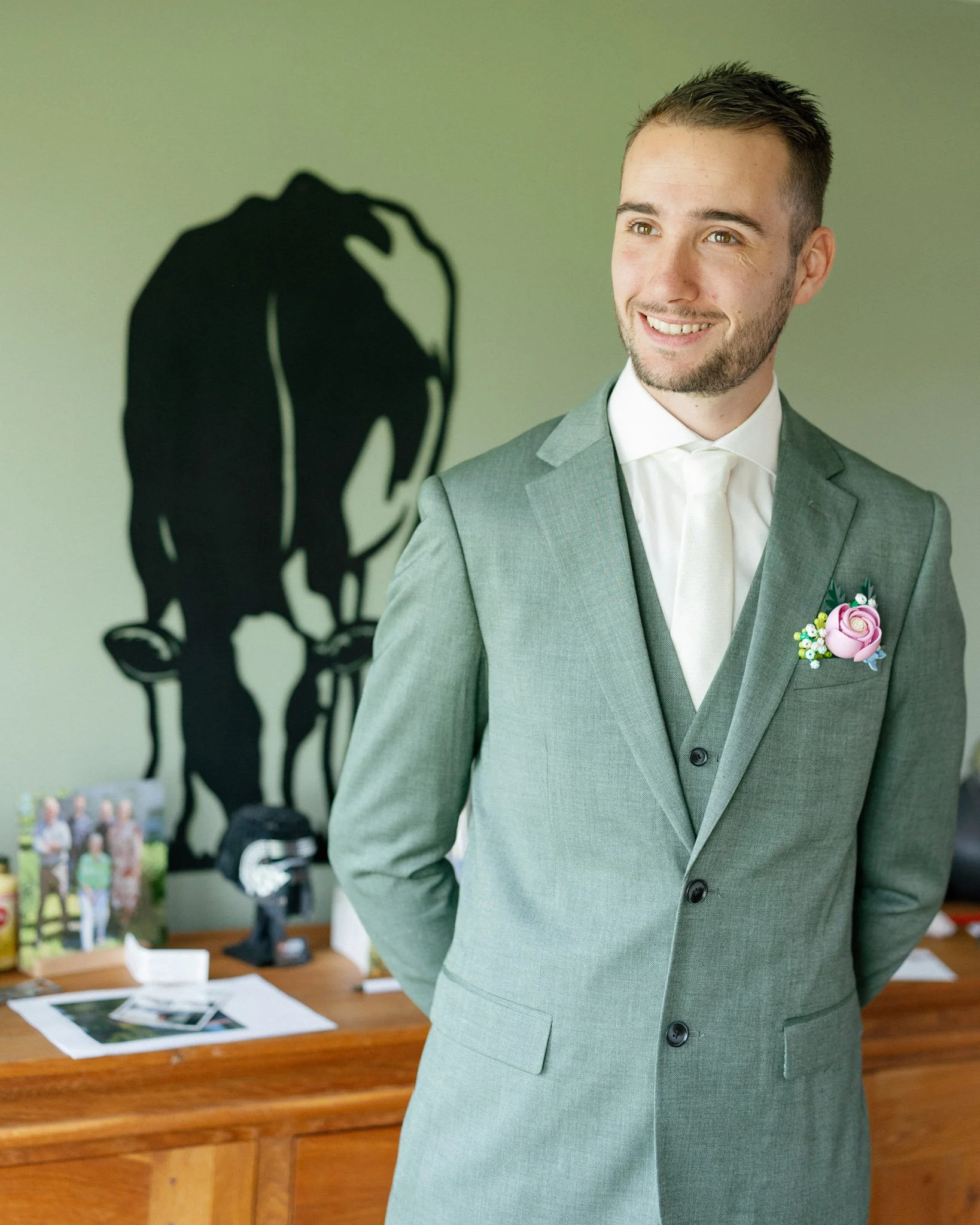 A young man in a gray suit and white shirt smiling, standing in front of a light green wall with a shadow of a horse on it. There is a wooden table with papers and photographs in the background.