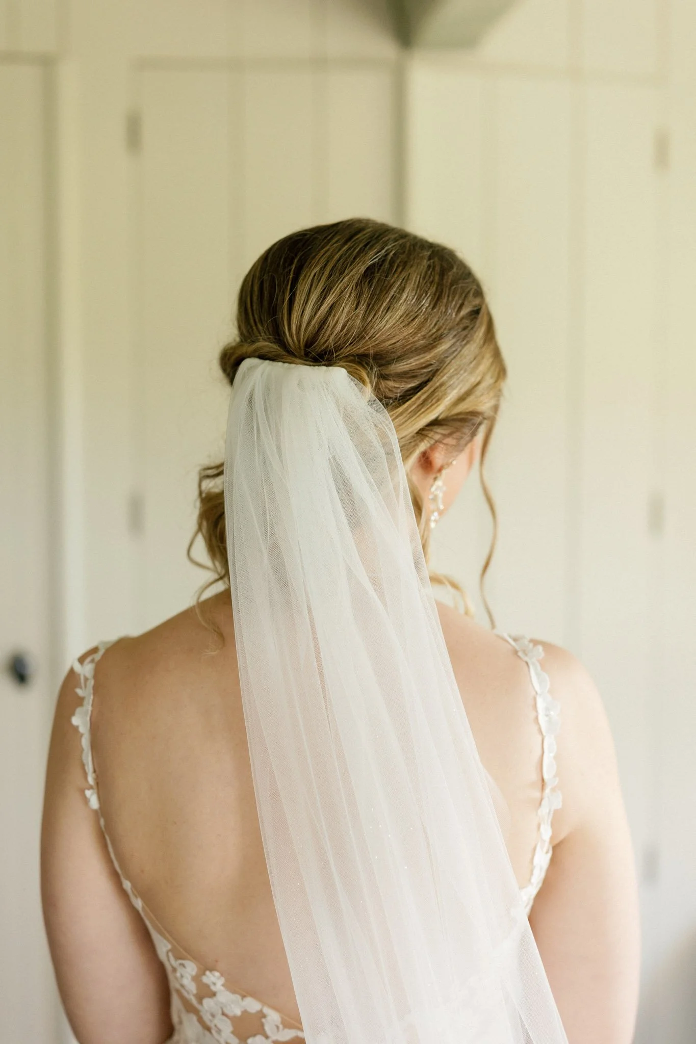 Back of a bride with styled hair, wearing a veil and a lace dress with floral details, facing away.