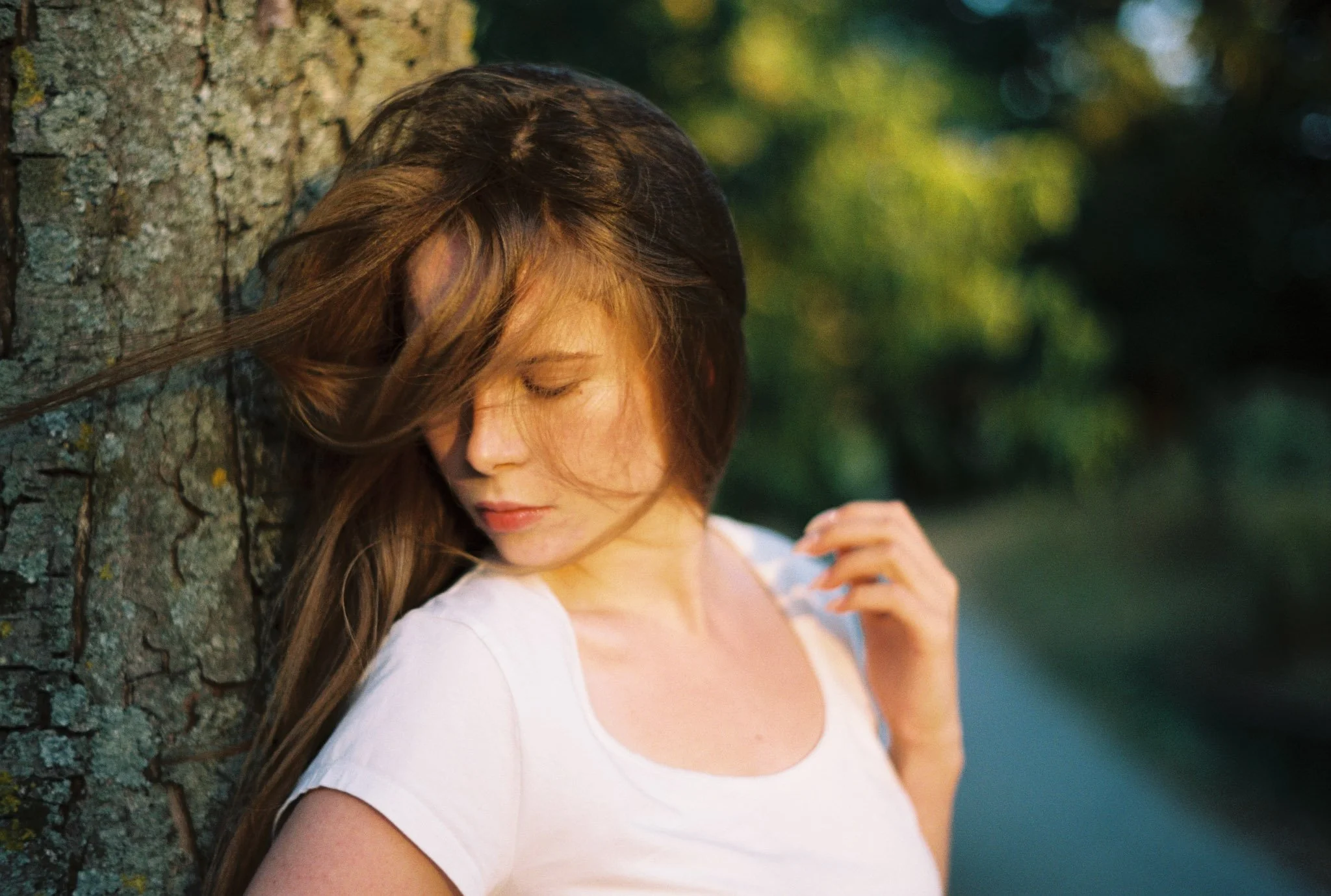 A woman with long brown hair leaning her head against a tree, with her eyes closed and a peaceful expression. She is wearing a white shirt and standing outdoors with a blurred green background.