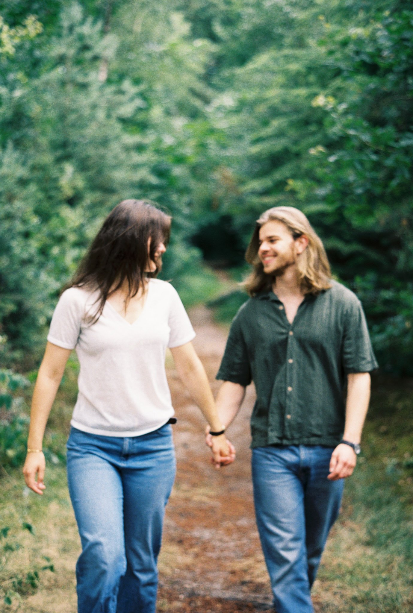 A couple holding hands and walking down a wooded trail on a sunny day, smiling at each other.