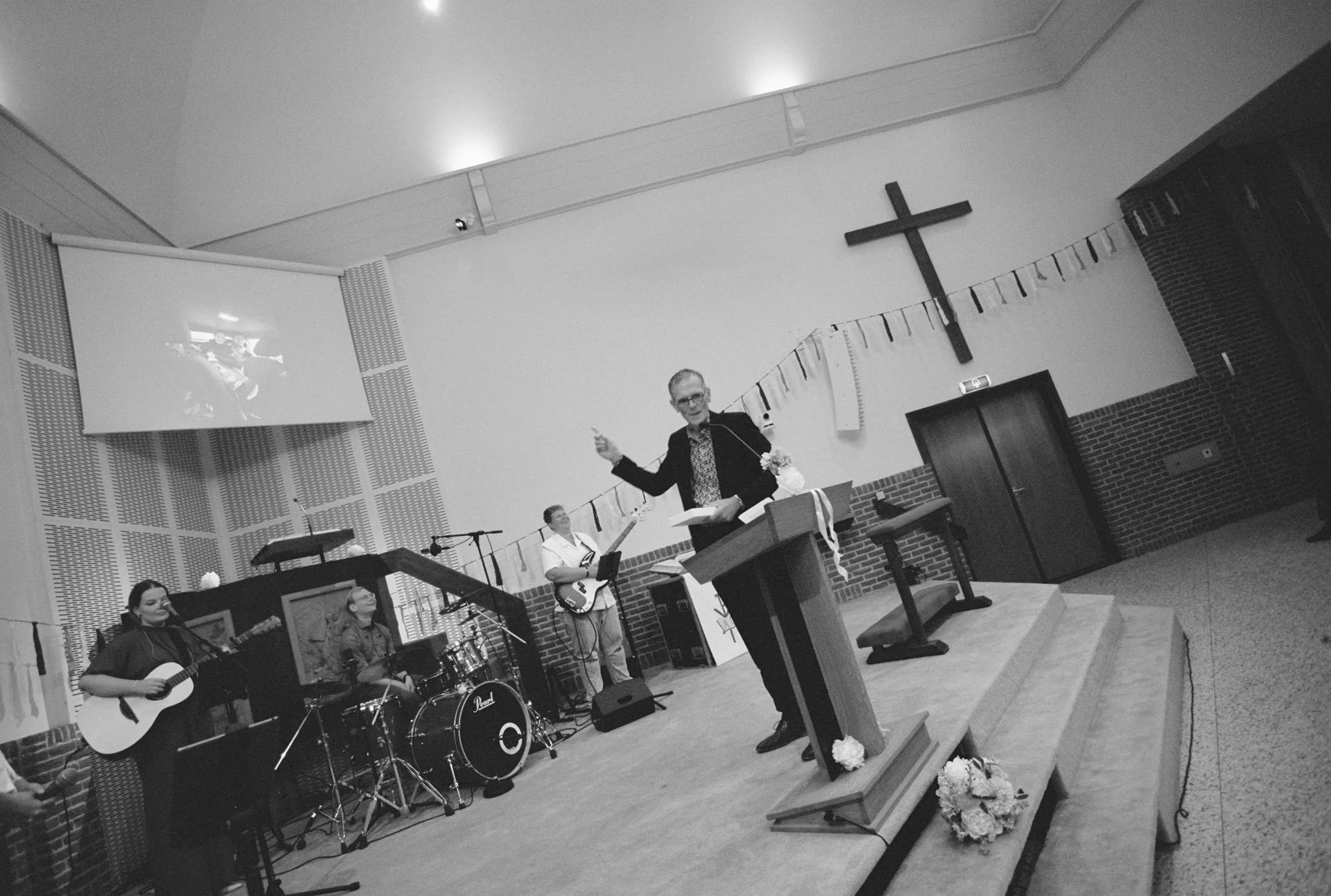 A church service with musicians and a speaker at the pulpit. The church interior features a large cross, a projector screen, and flower decorations.