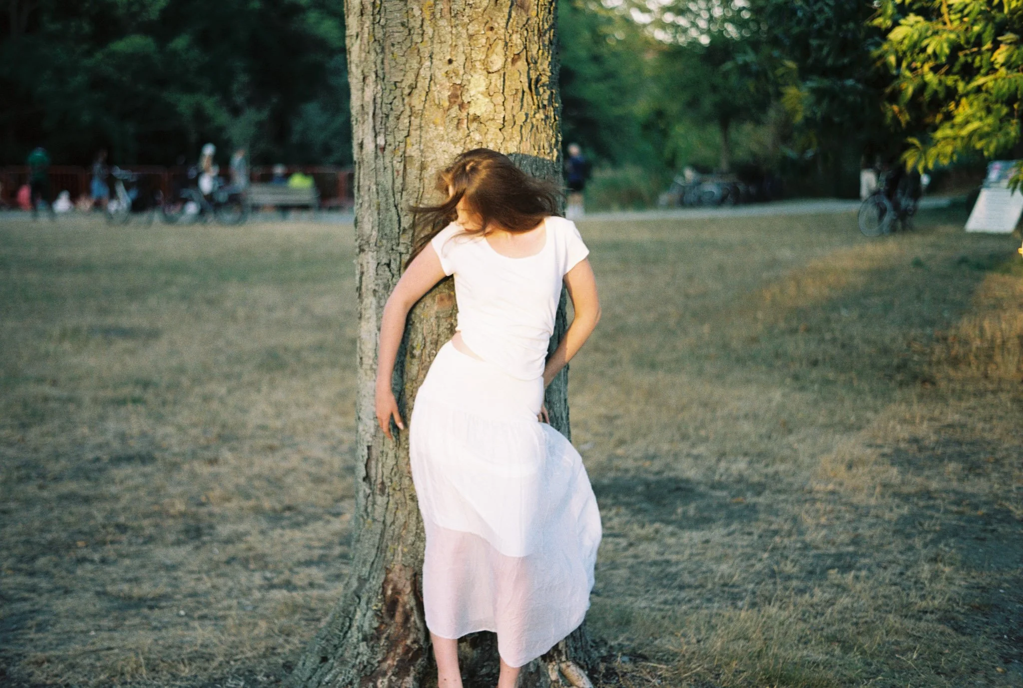 A woman with long brown hair in a white dress is leaning against a tree in a park, with others in the background.