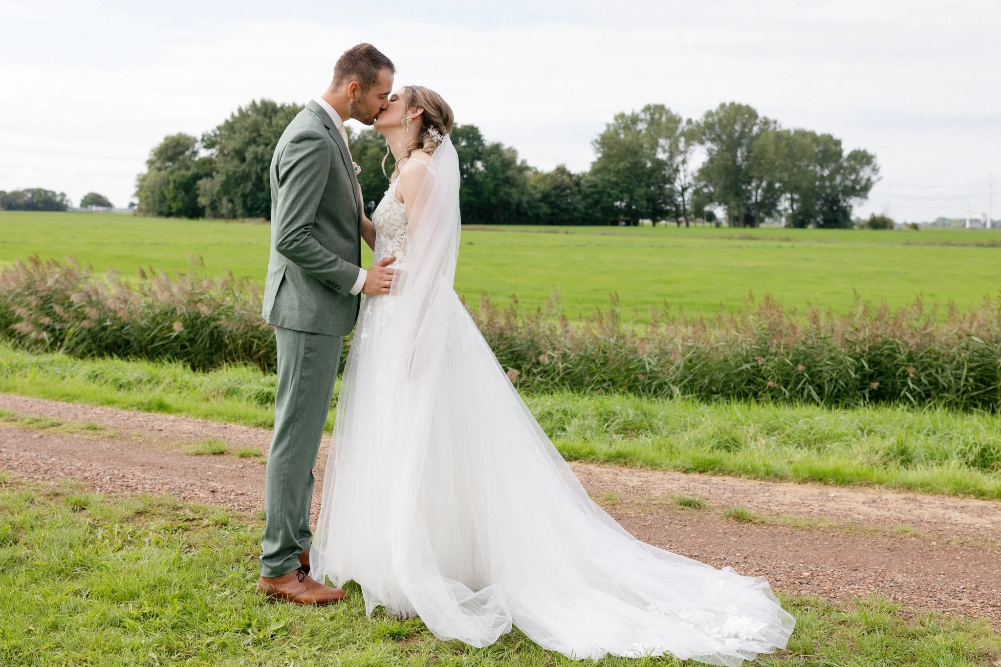 A bride and groom sharing a kiss outdoors during their wedding, with green fields and trees in the background.