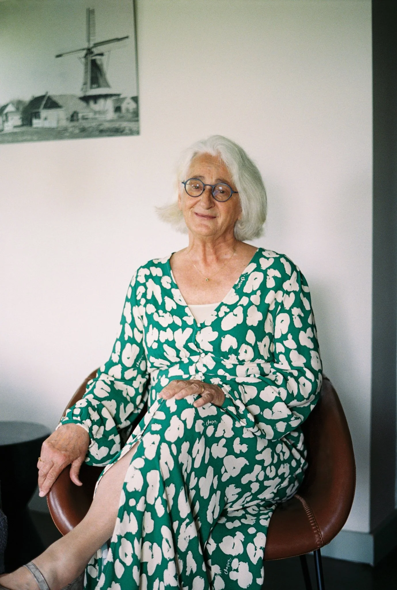 An elderly woman with white hair and glasses sitting in a brown chair. She is wearing a green dress with white abstract patterns. Someone's foot is visible in the foreground, touching her foot.