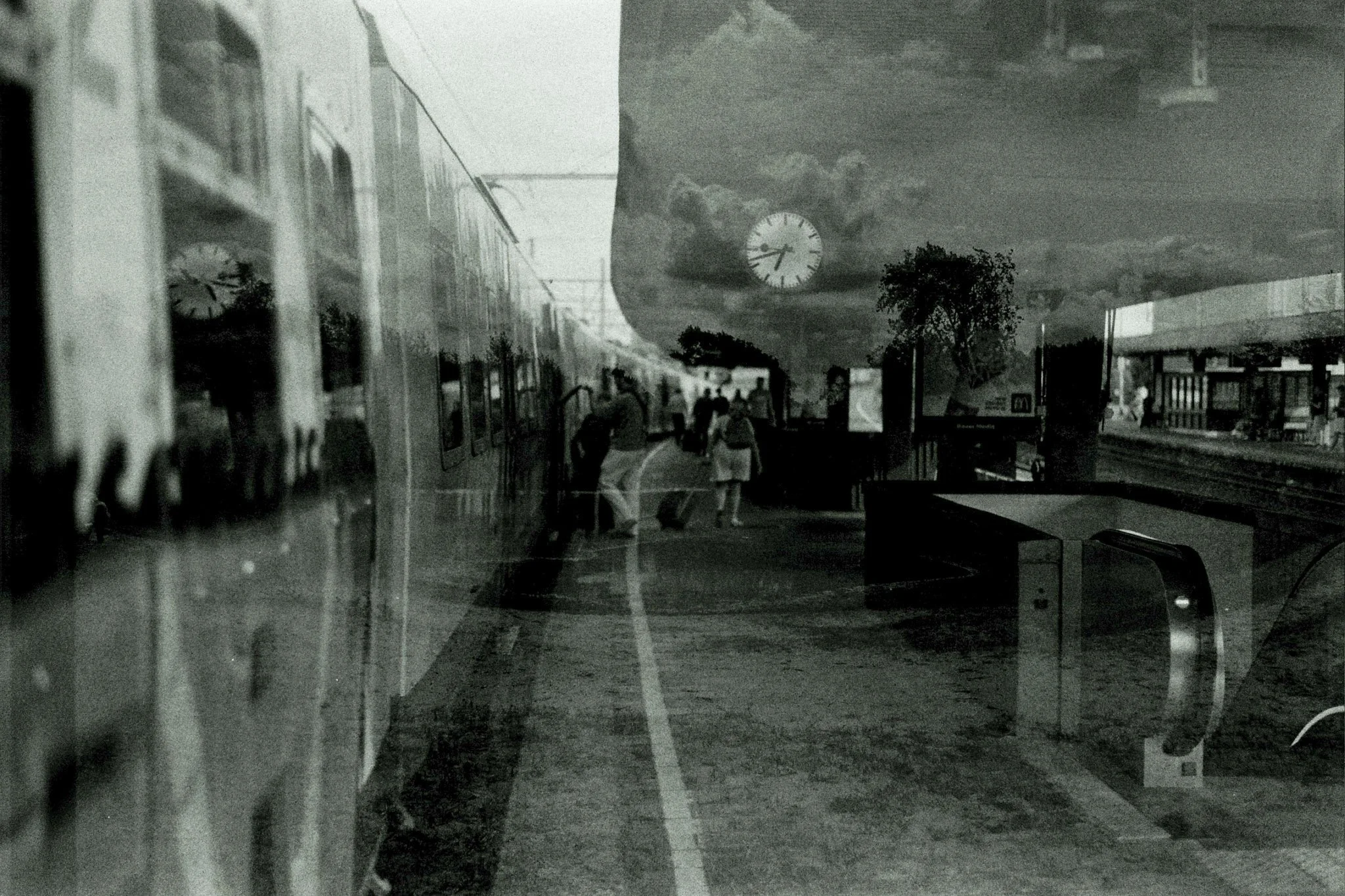 Black and white photo of a train station platform with people waiting, a train, a clock showing 7:05, trees, and buildings in the background.