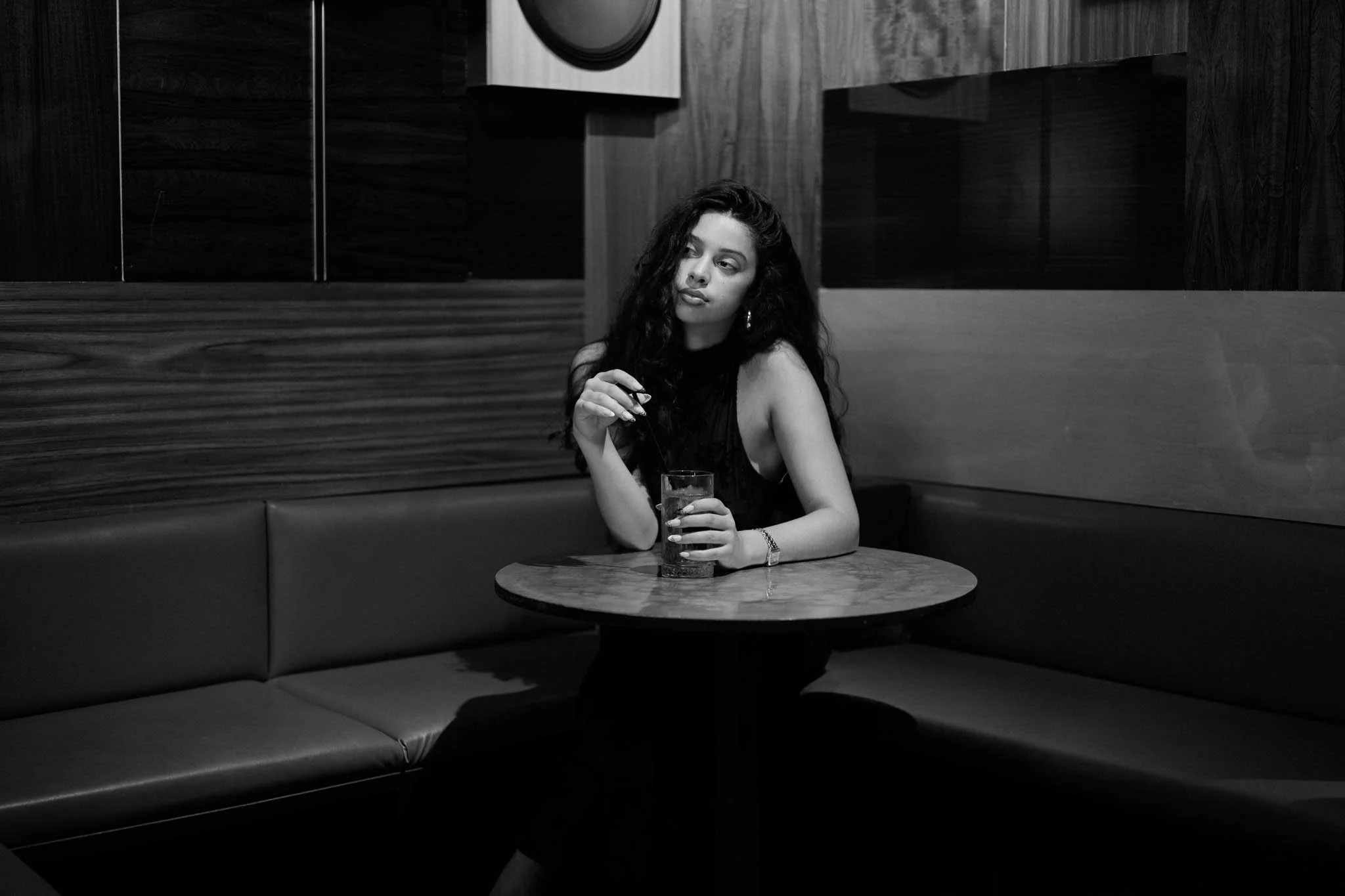 A woman with long, curly hair and earrings is sitting alone at a round table, holding a drink in one hand, in a dimly lit restaurant with dark wood paneling and booth seating.