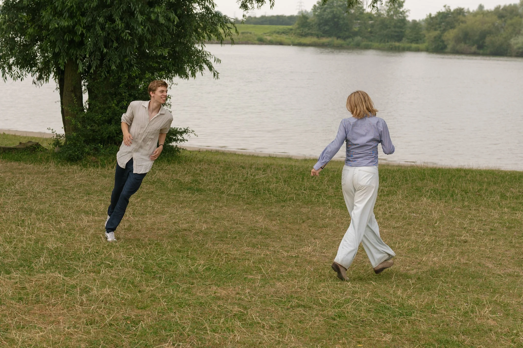 Two young adults, a man and a woman, are outdoors near a lake or river, walking and laughing beneath a large tree on a grassy area.