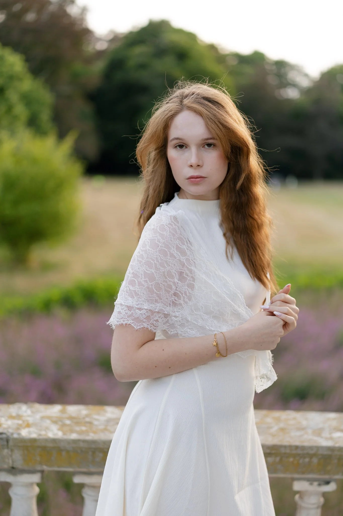 A young woman with long, wavy red hair wearing a white dress with lace details on the sleeves, standing outdoors near a balcony with stone balustrades, in front of a background of trees and greenery, possibly during dusk or dawn.