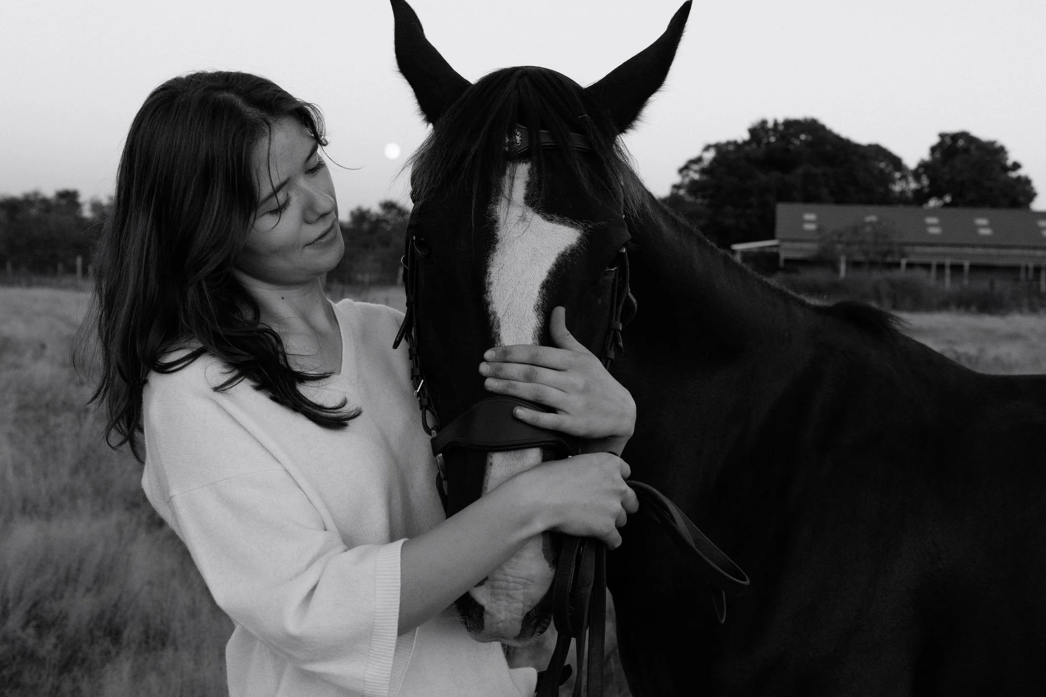 A woman with long hair wearing a light-colored sweater holding a horse's head, with her hand on the horse's face, in an outdoor rural setting with trees and a barn in the background.