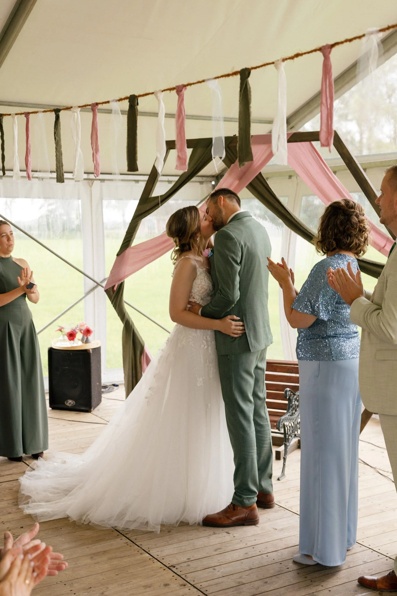 A bride and groom share a kiss during their wedding ceremony under a fabric-draped arch with pink and green accents, while guests clap and celebrate.