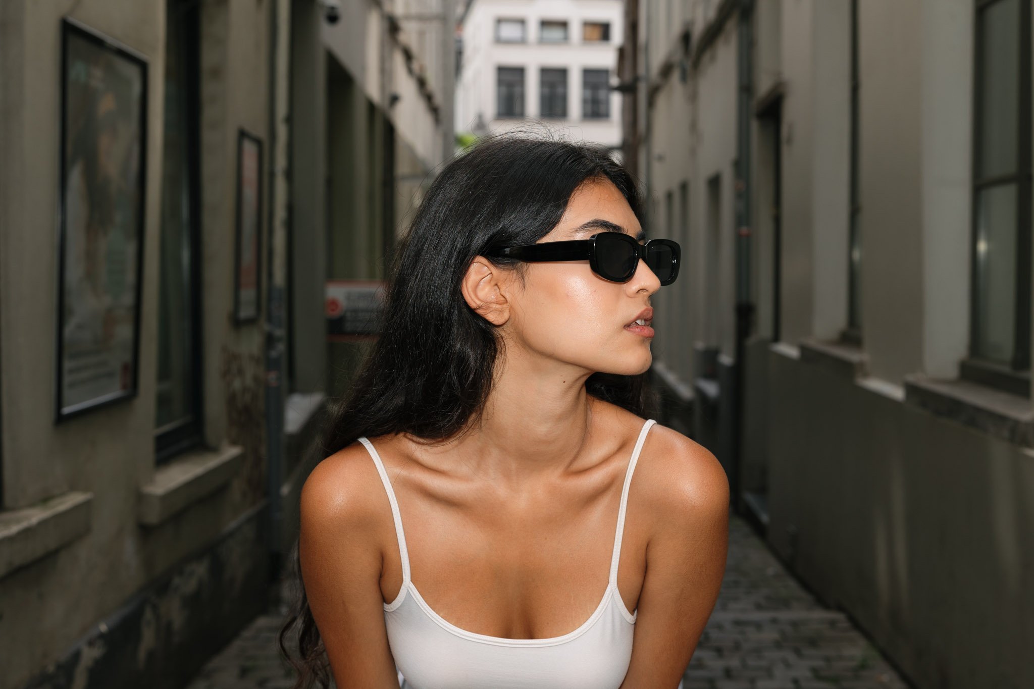 A woman with long dark hair wearing black sunglasses and a white tank top standing in an alleyway between buildings.