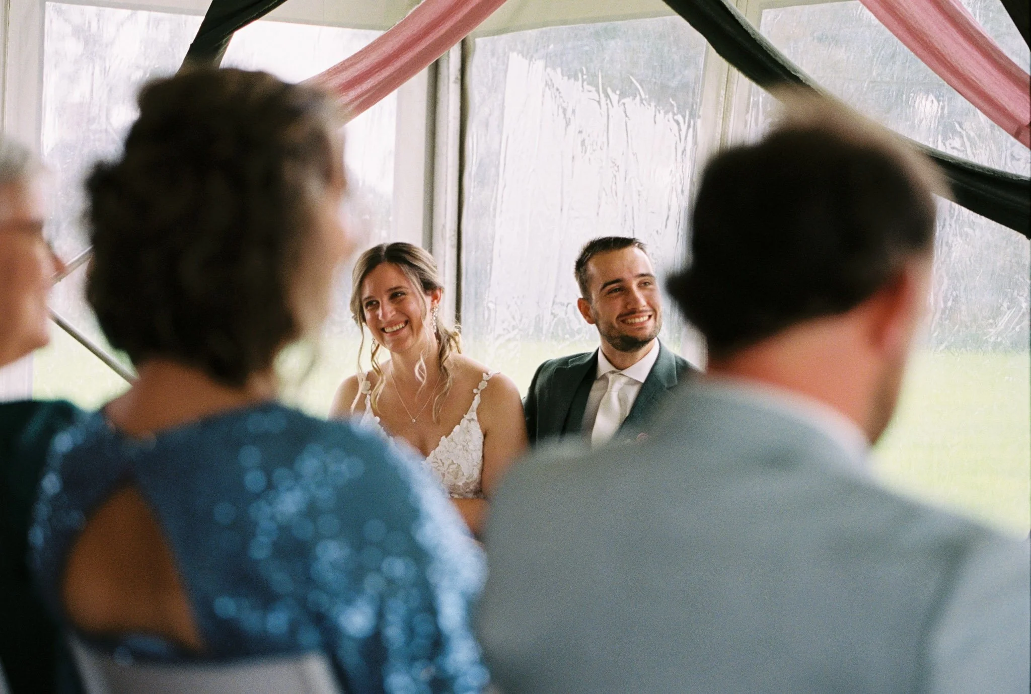 A group of people at a wedding reception, with the bride and groom smiling in the background, seen through the blurred heads and shoulders of guests in the foreground inside a tent.