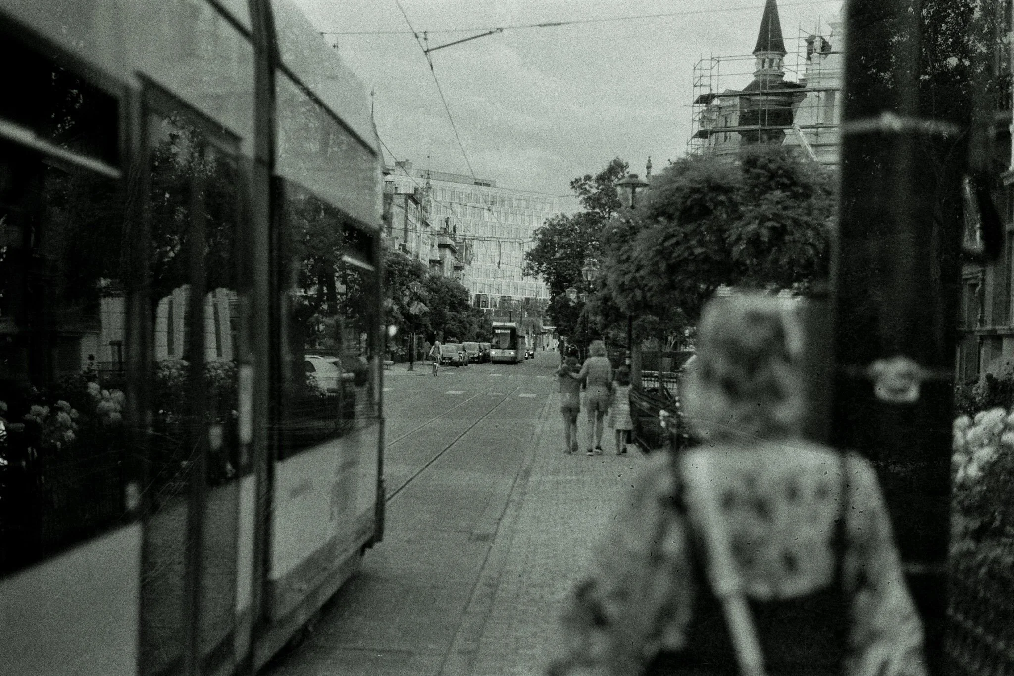 A street scene viewed through a glass window, with reflections on the glass. There are people walking on the sidewalk, and a tram or bus is visible on the street. The background features trees and a building with scaffolding around a tower.