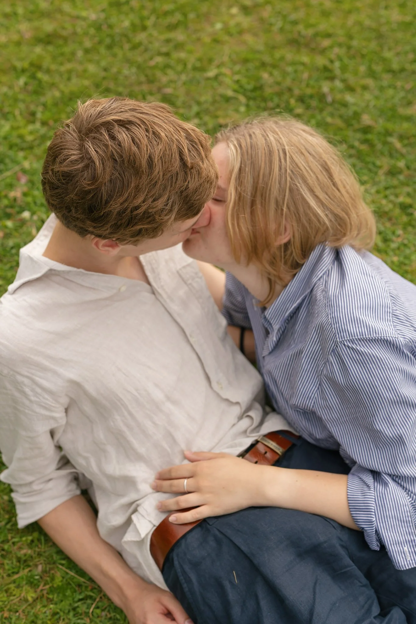 A couple kissing while sitting on grass, with the woman resting her hand on the man's stomach.