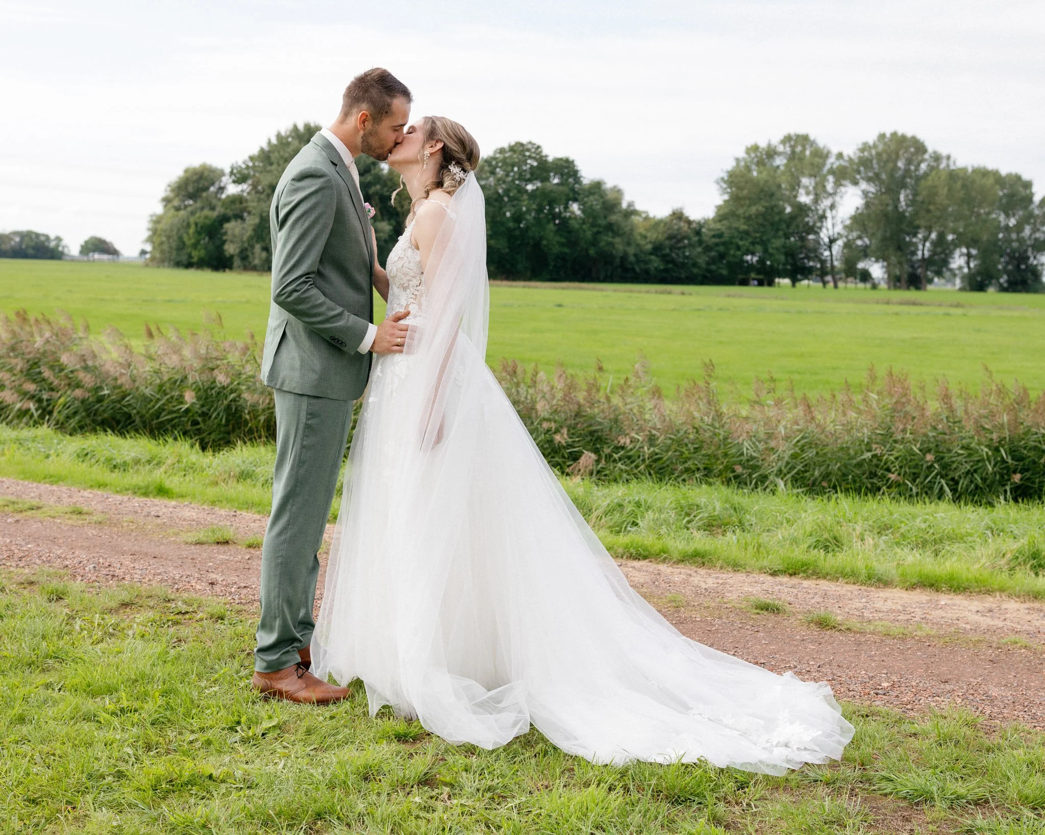 A couple in wedding attire sharing a kiss in an outdoor setting with green grass, trees, and cloudy sky.