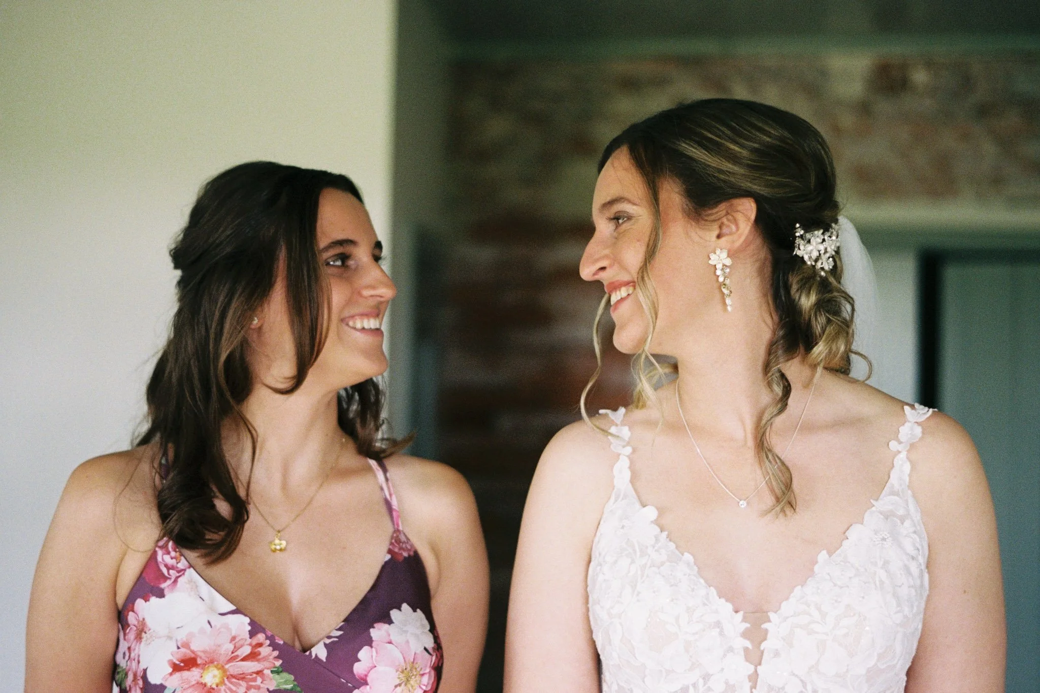 Two women in wedding dresses smiling at each other indoors.