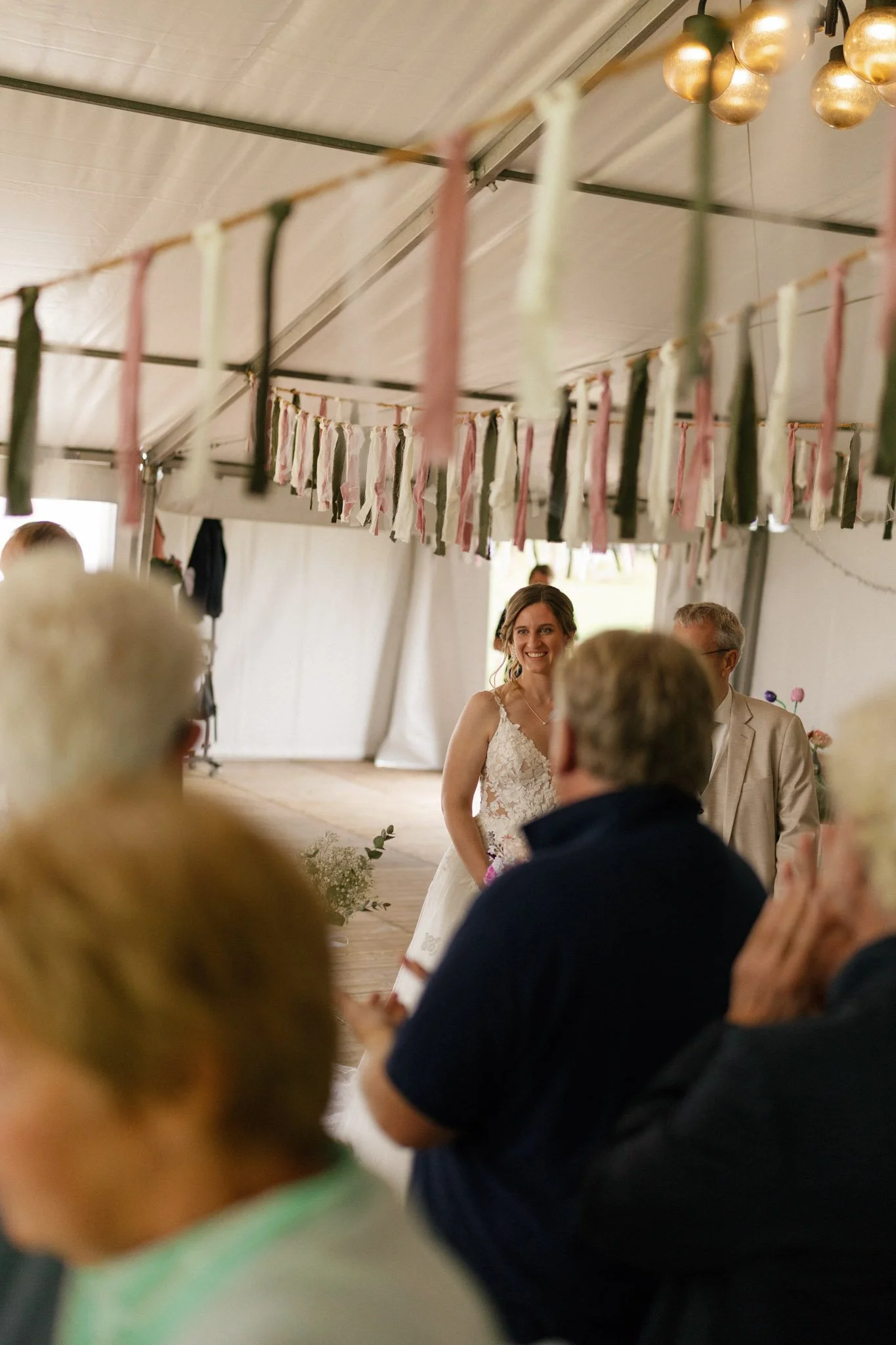 A wedding celebration inside a tent with a bride in a white lace wedding dress and a man in a beige suit. Decorations include hanging fabric strips and golden hanging lights. Several guests are present, some clapping and engaged in the moment.