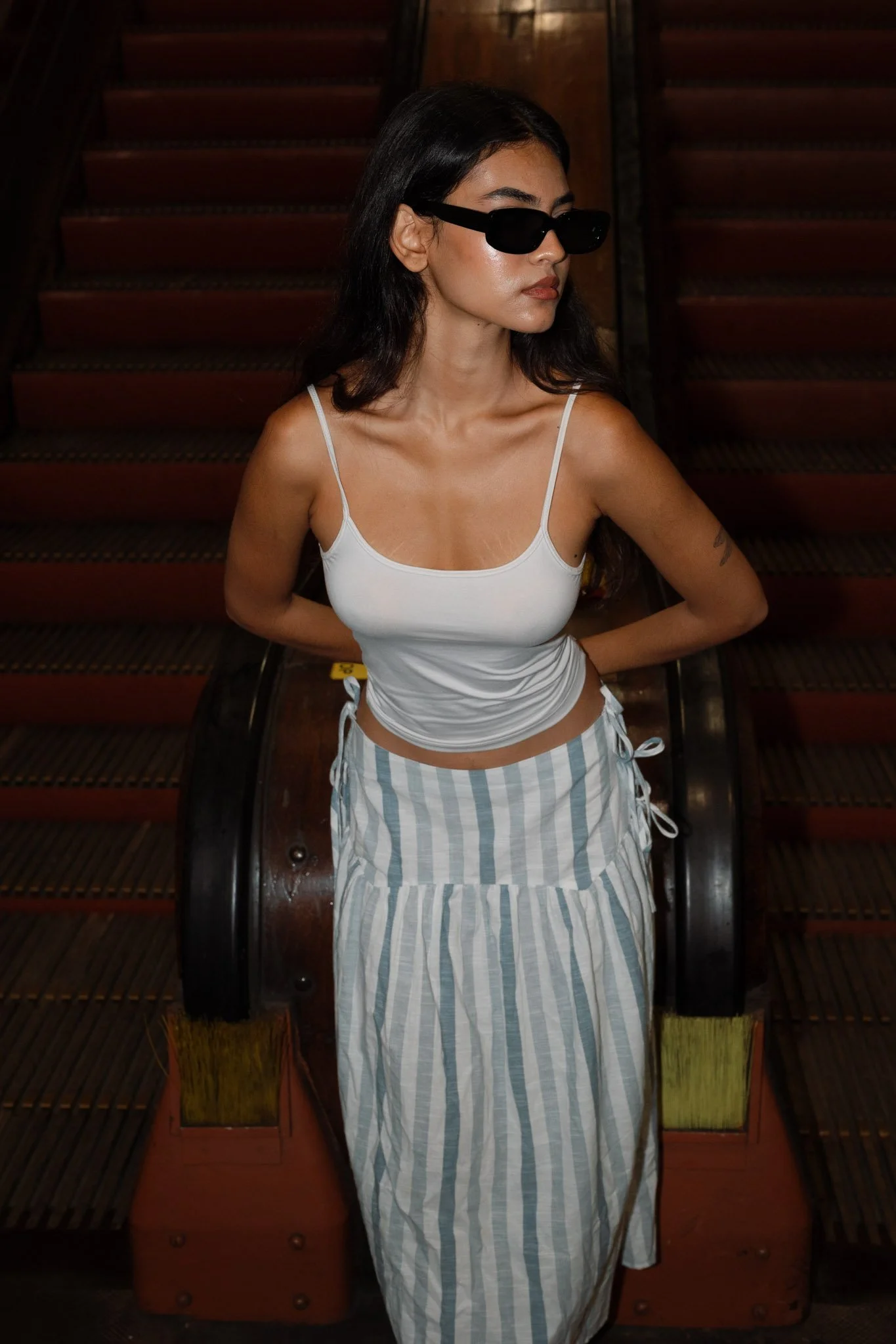 A woman with long dark hair wearing black sunglasses, a white spaghetti strap top, and a striped skirt standing in front of a vintage wooden escalator in a dimly lit indoor setting.