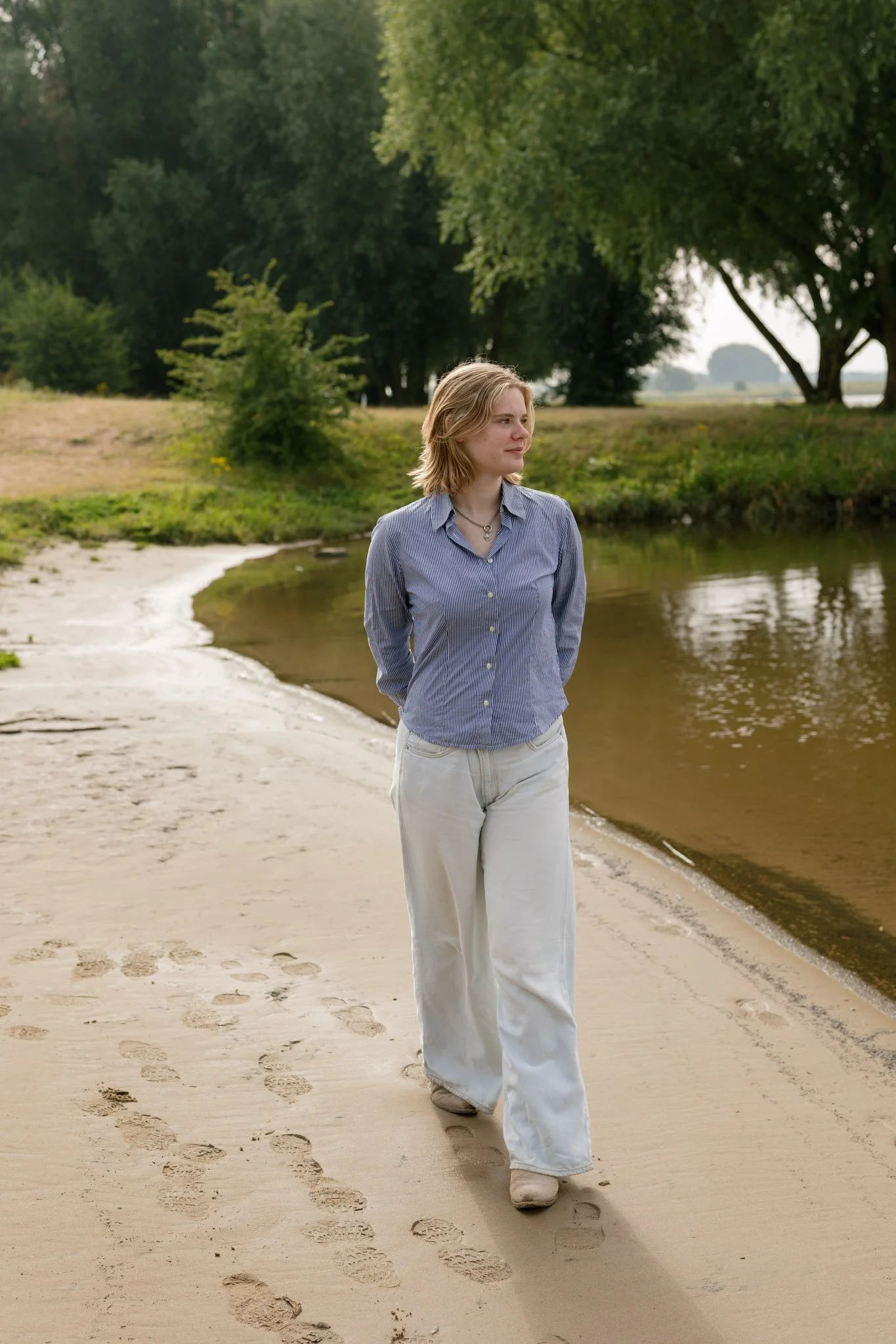 A woman walking along the sandy beach near a river, with footprints behind her and trees in the background.