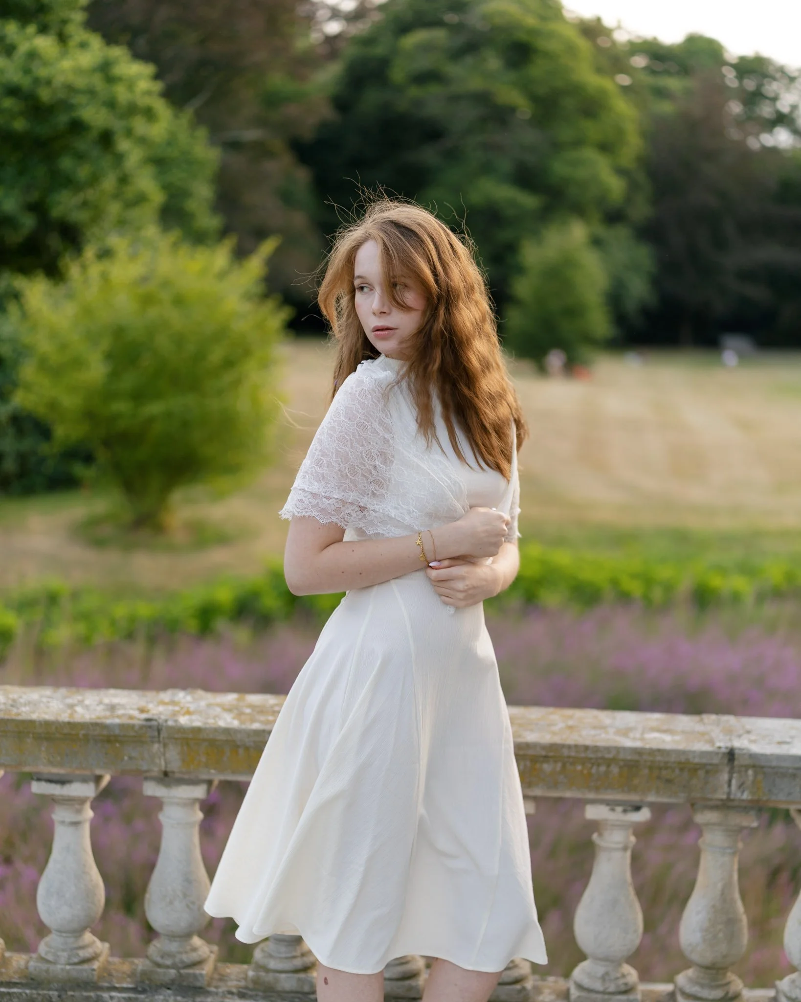 A young woman with long red hair in a white lace dress standing outdoors near a stone railing, with green trees and purple flowers in the background.